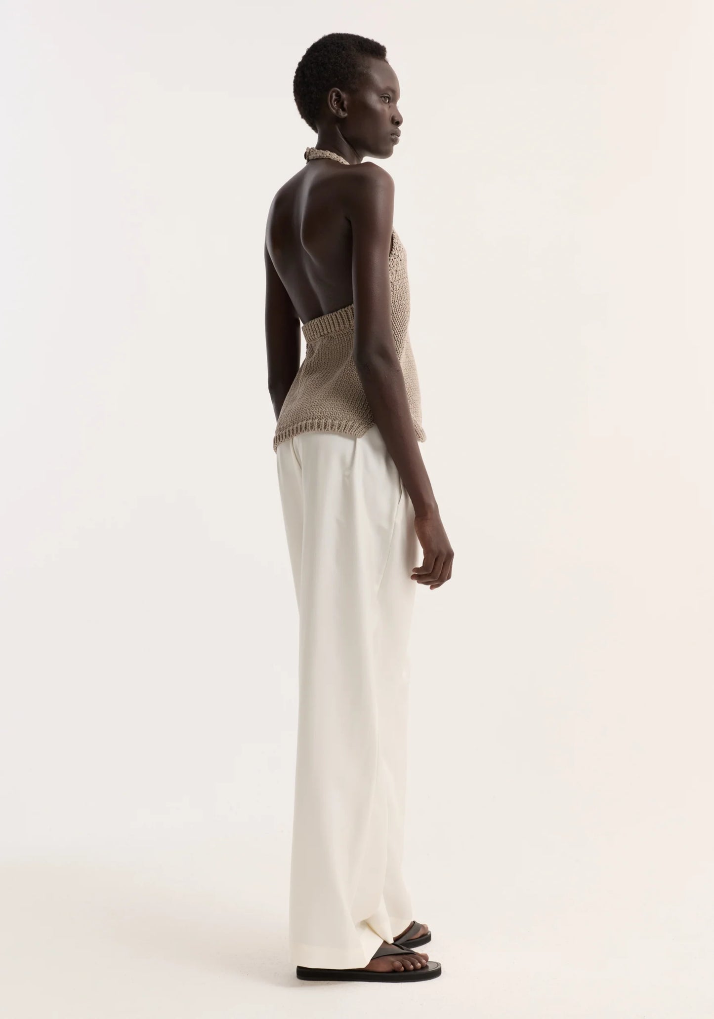 Woman wearing a beige sleeveless top and white pants on a plain background