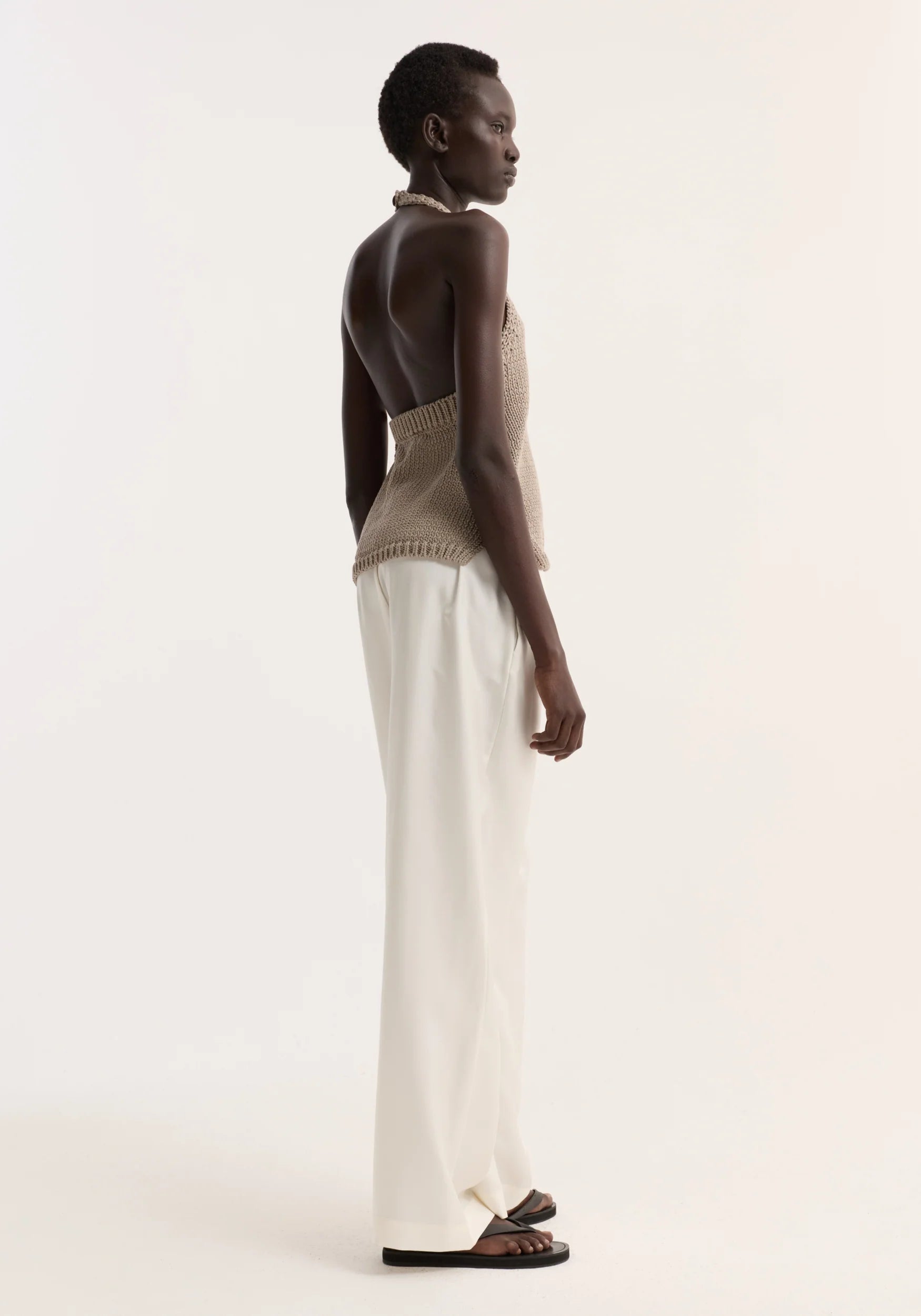 Woman wearing a beige sleeveless top and white pants on a plain background
