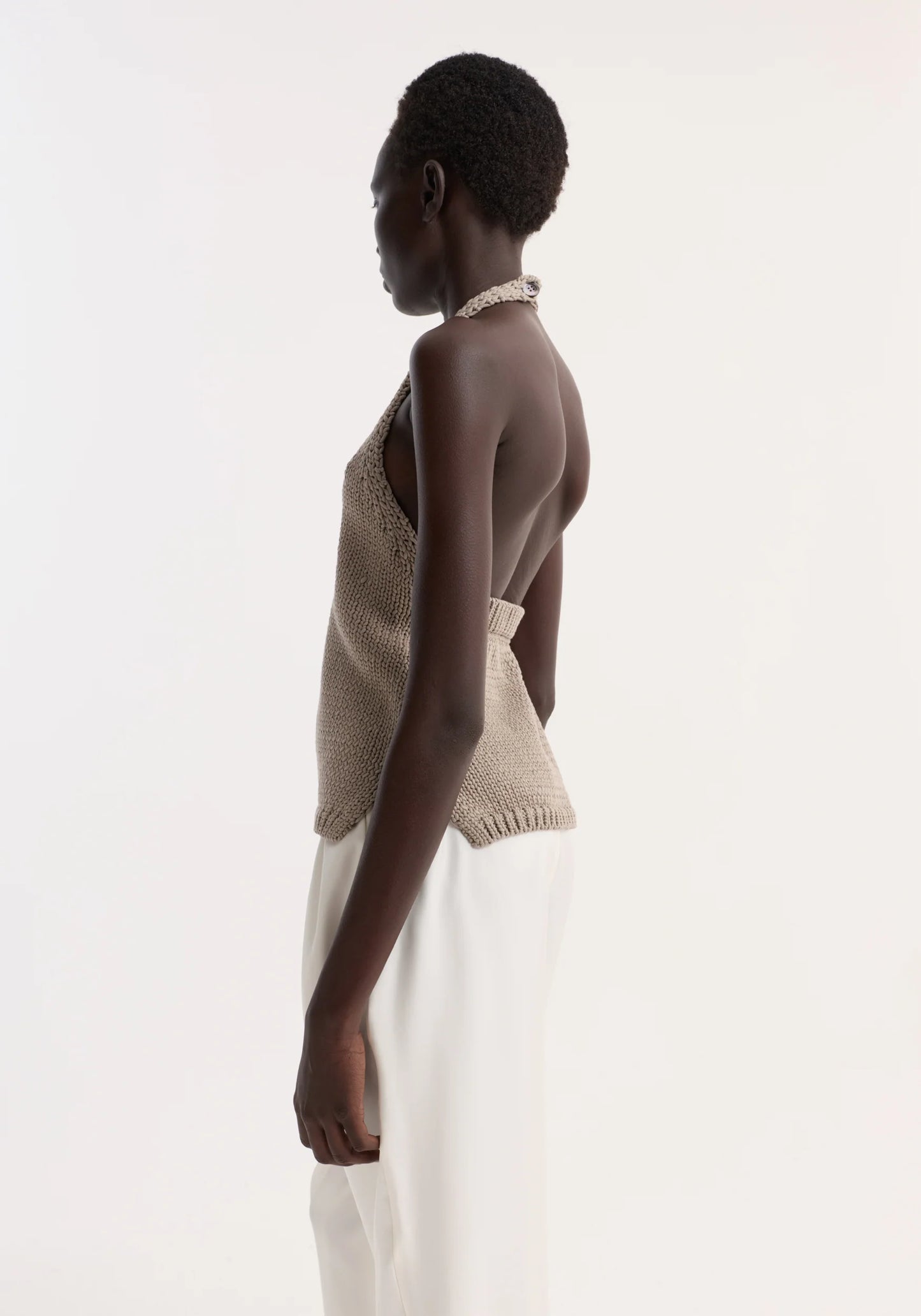 Woman wearing a beige knitted top and white pants on a plain background