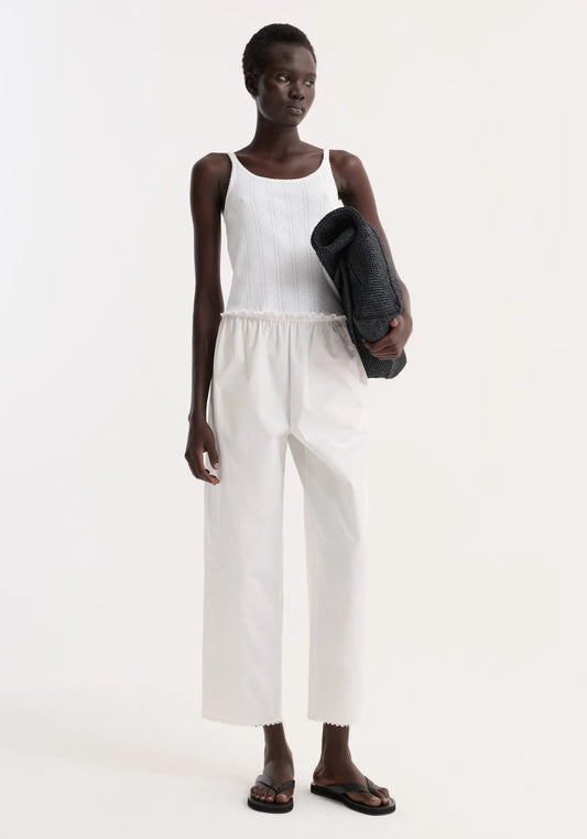 Person wearing a white sleeveless top and white pants holding a black yoga mat on a white background