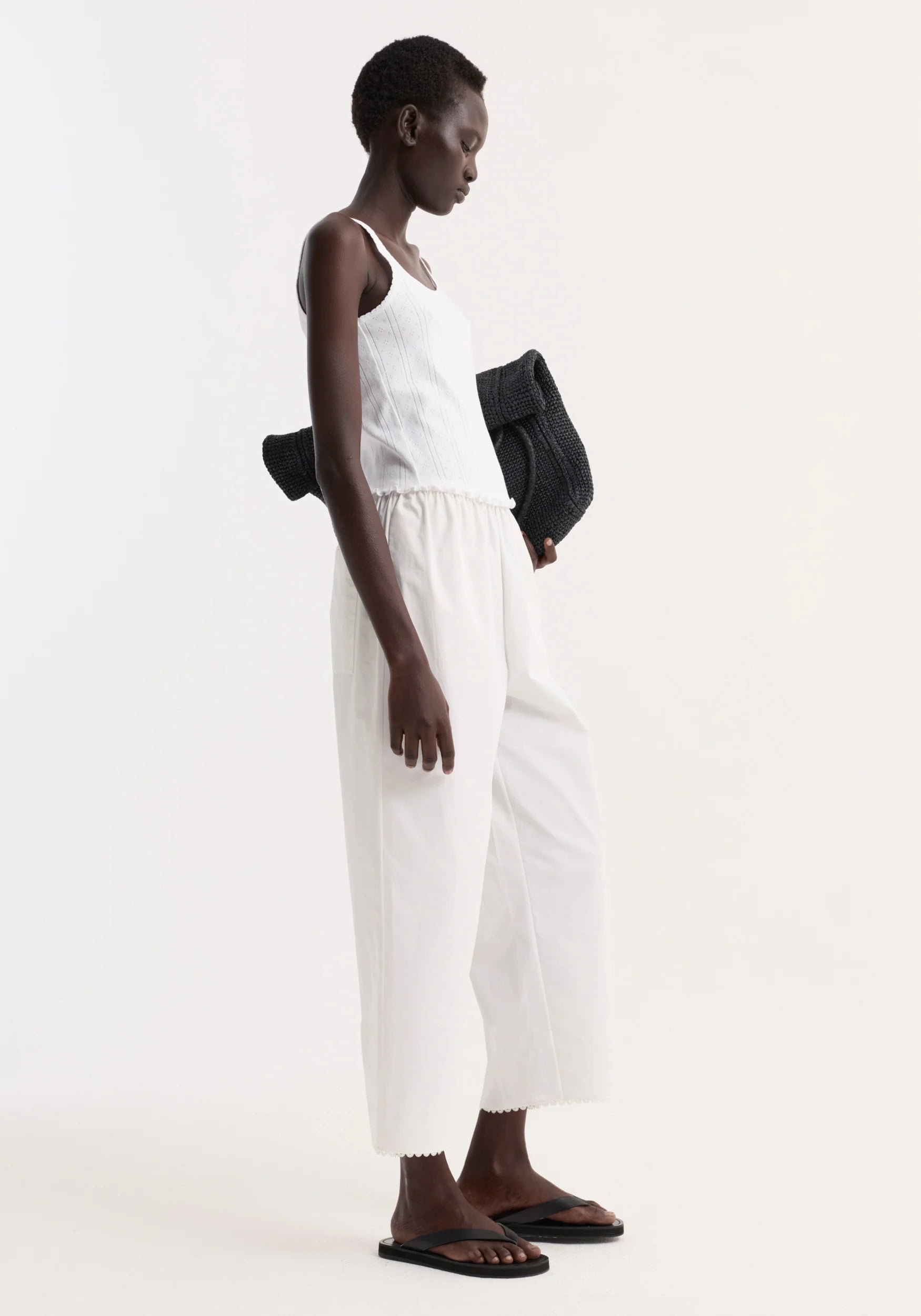 Person wearing a white sleeveless top and pants with a black bag on a white background