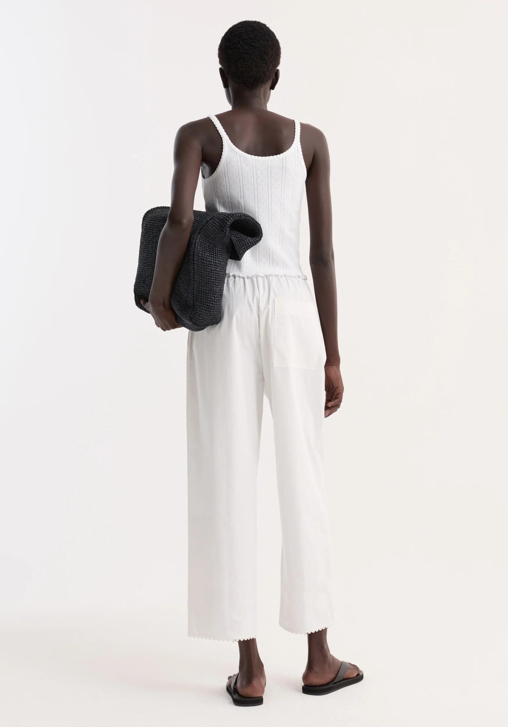 Person wearing a white tank top and wide-leg pants holding a black jacket on a white background