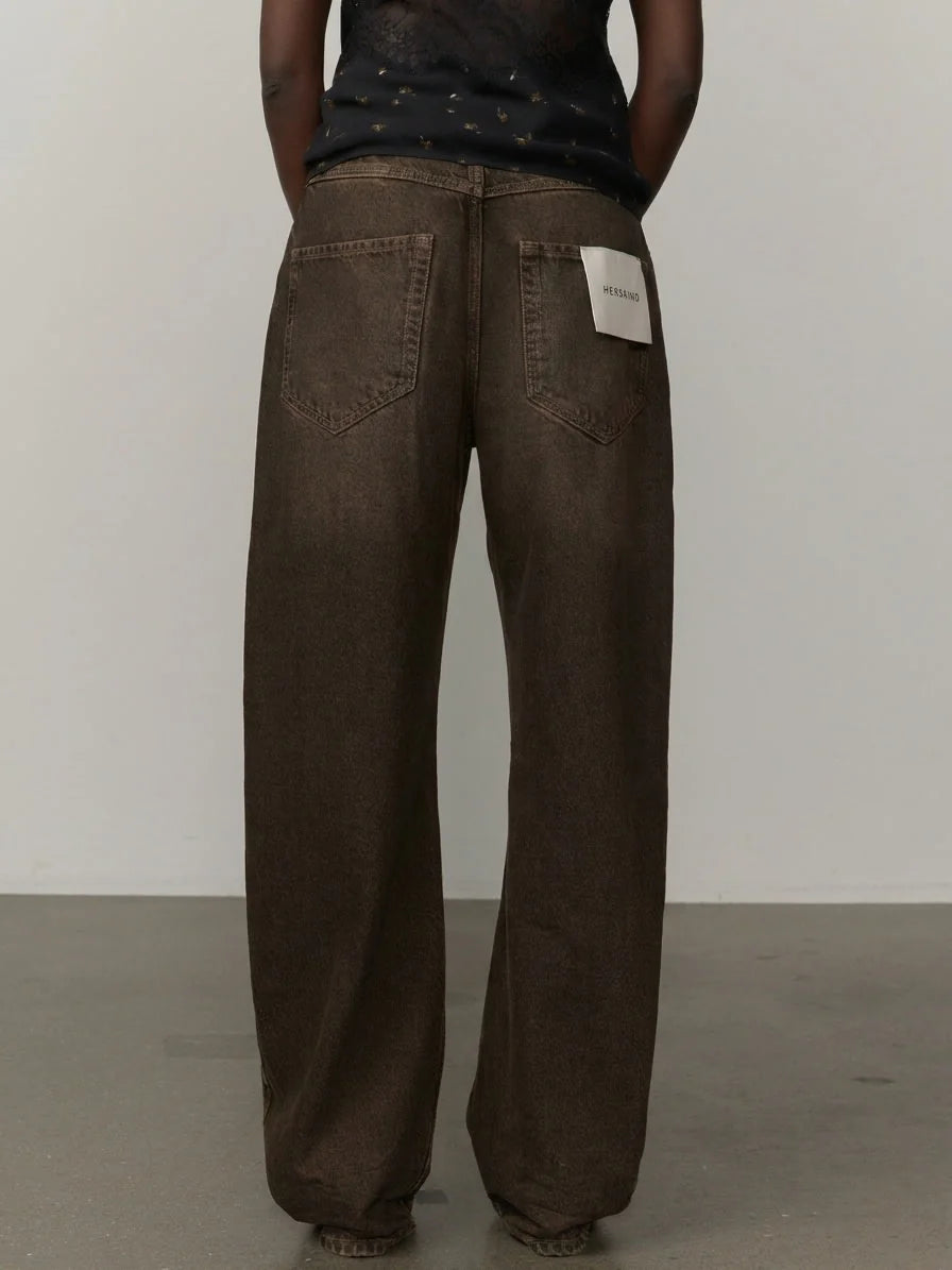A person shown from the back wearing very loose, wide-leg brown pants with a white brand patch on the back right pocket, against a plain light gray wall and concrete floor.