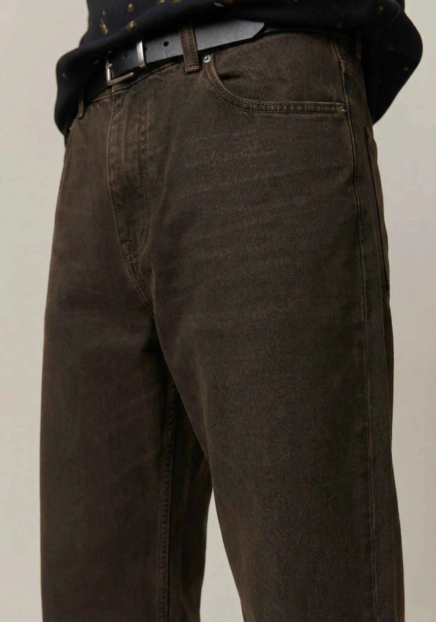 Close-up of a person wearing dark brown washed denim jeans with a black belt and a dark patterned shirt, shown from the waist down against a plain light background.