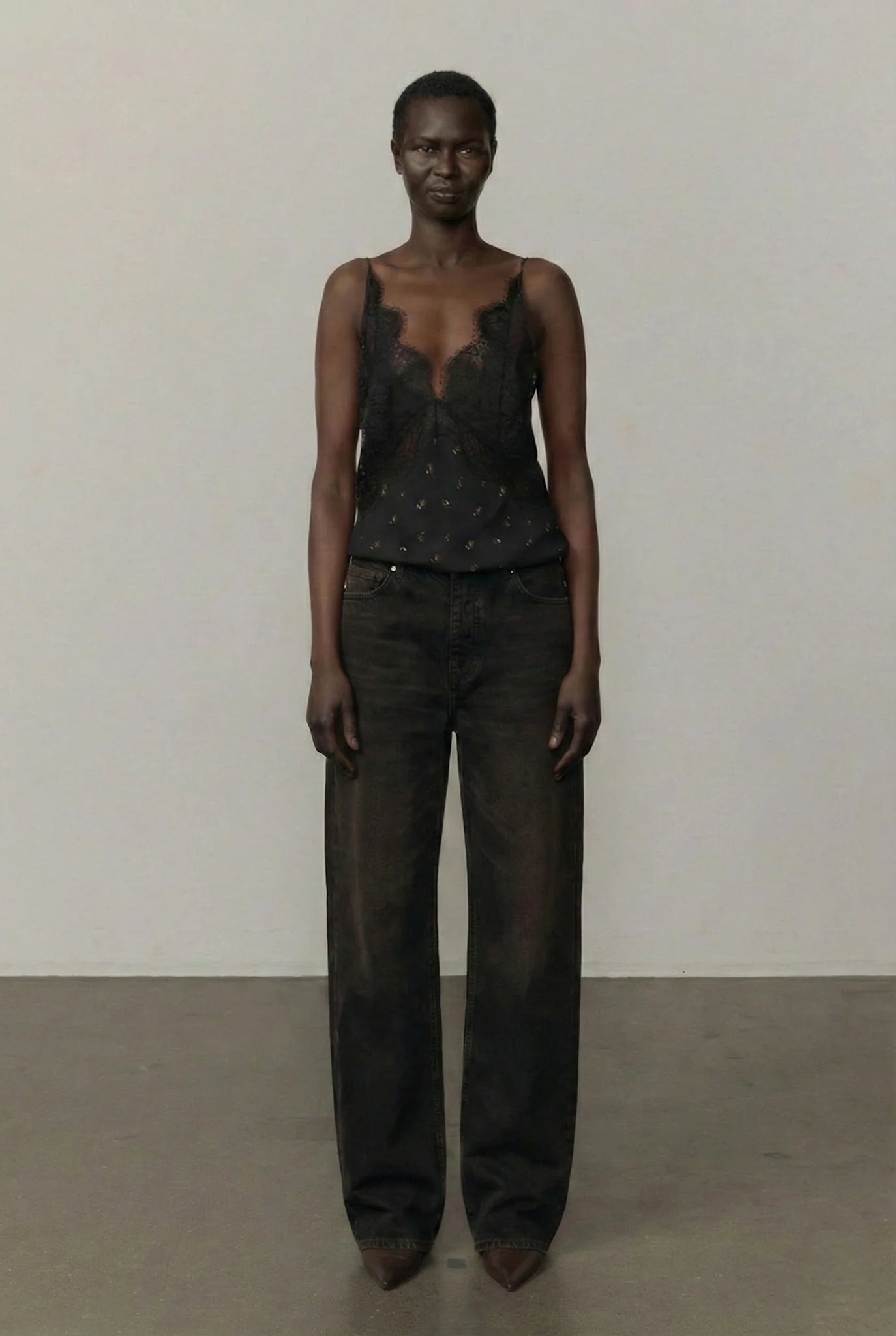 A model stands facing forward in a minimalist studio setting, wearing a sheer black lace camisole top with small metallic embellishments and dark wide-leg jeans.