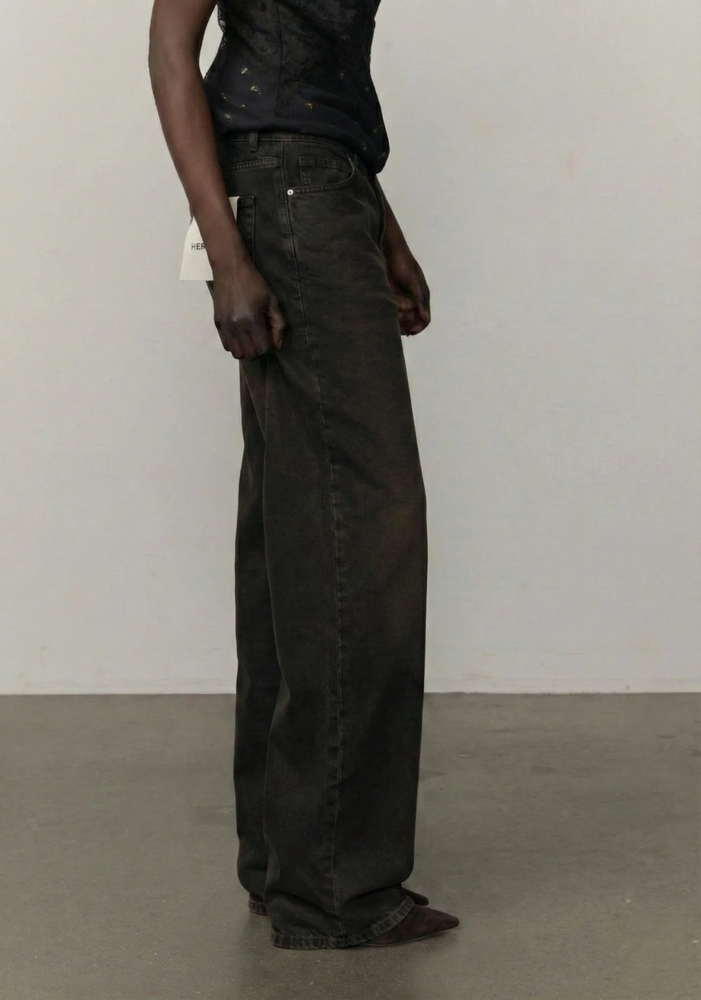 A person wearing very wide-leg dark charcoal gray or black denim jeans, paired with a dark top, standing in a minimalist studio with a white wall and gray floor.