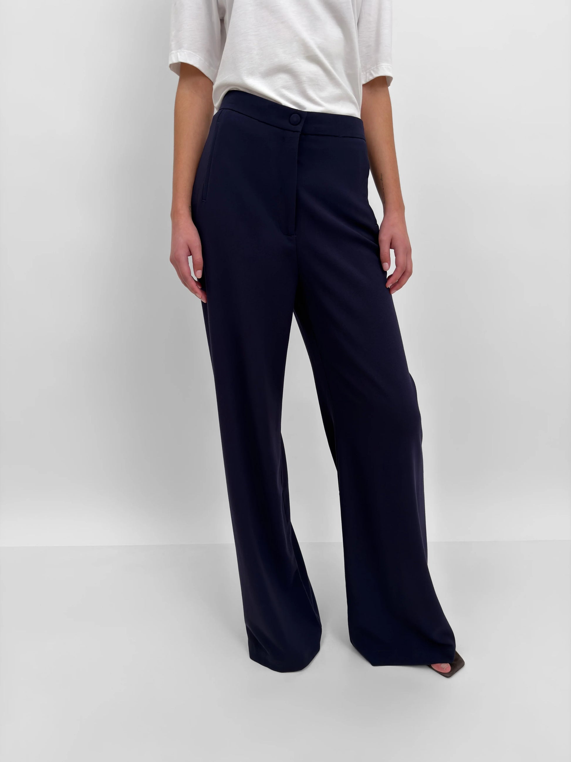 A model wearing dark navy wide-leg trousers and a white short-sleeve top, standing against a light gray studio background.