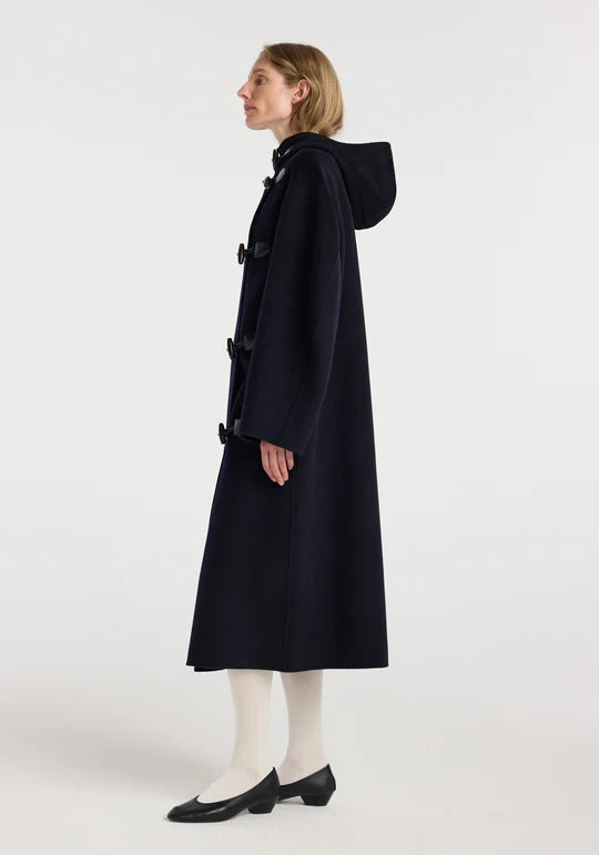 A woman modeling a long navy hooded duffle coat with large dark buttons, paired with white tights and black shoes, shown in profile against a plain light gray studio background.