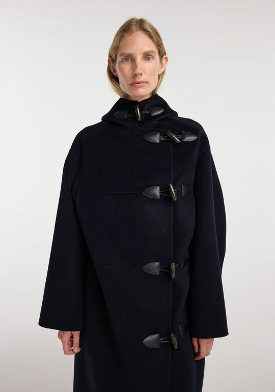 A woman modeling a dark navy hooded duffle coat with dark toggle closures against a plain light gray studio background.