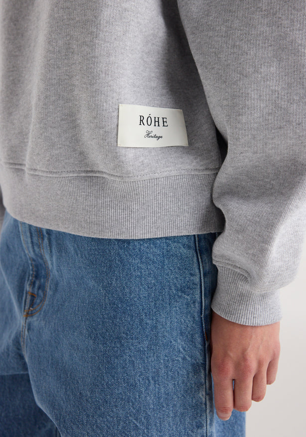 Gray sweatshirt with a brand label on the chest, worn by a person with blue jeans.