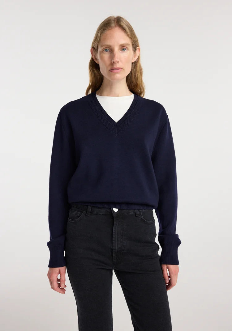 A woman modeling a navy blue V-neck knit sweater layered over a white crew-neck top and paired with black jeans, shot against a plain light gray studio background.