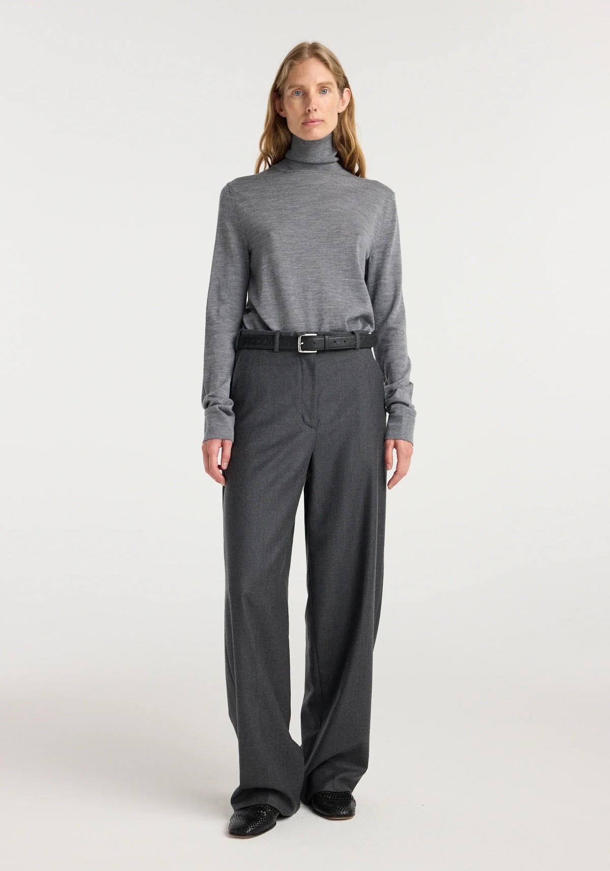 A woman stands against a light gray studio background wearing a gray long-sleeve turtleneck sweater, dark charcoal wide-leg trousers, a black belt, and black shoes.