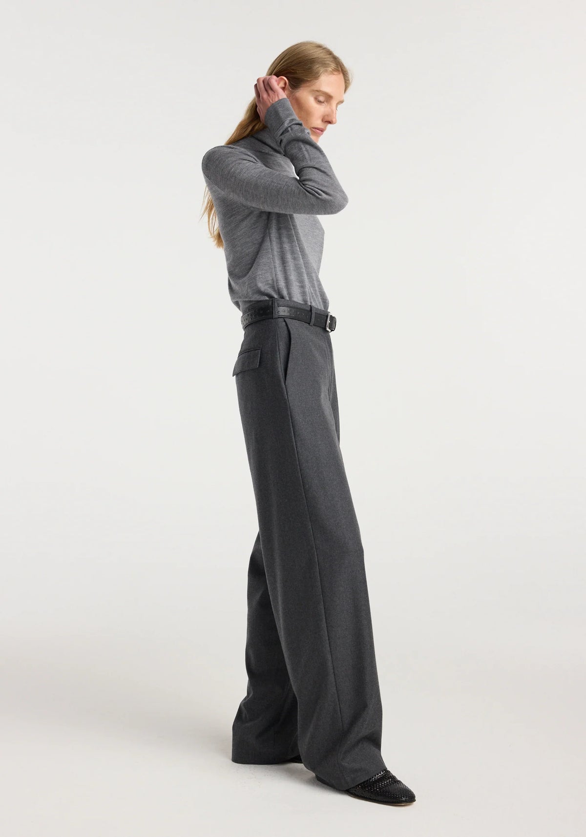 A woman stands in profile wearing a gray long-sleeve knit top and dark charcoal wide-leg trousers with a black belt, against a plain light gray studio background.