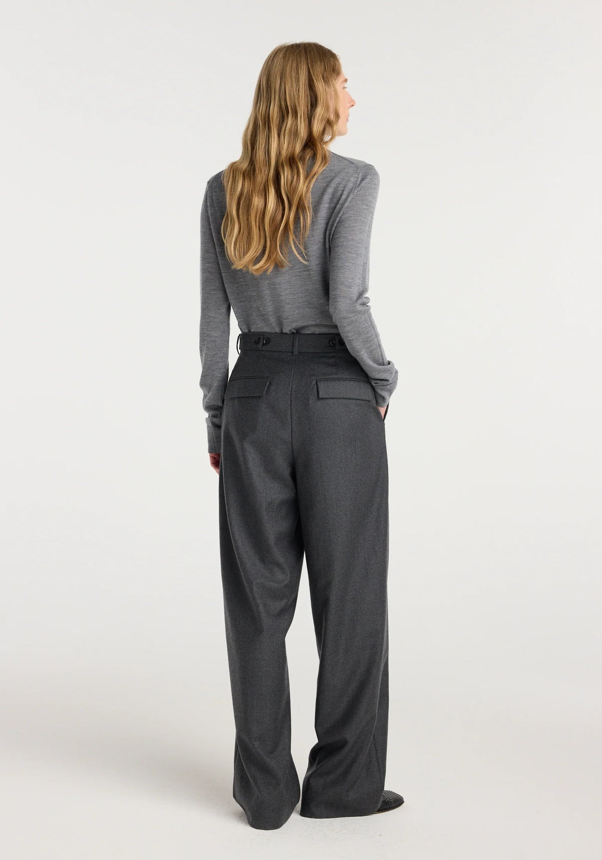 A woman shown from the back wearing a gray long-sleeve top and dark charcoal gray wide-leg trousers, standing against a plain light gray studio background.