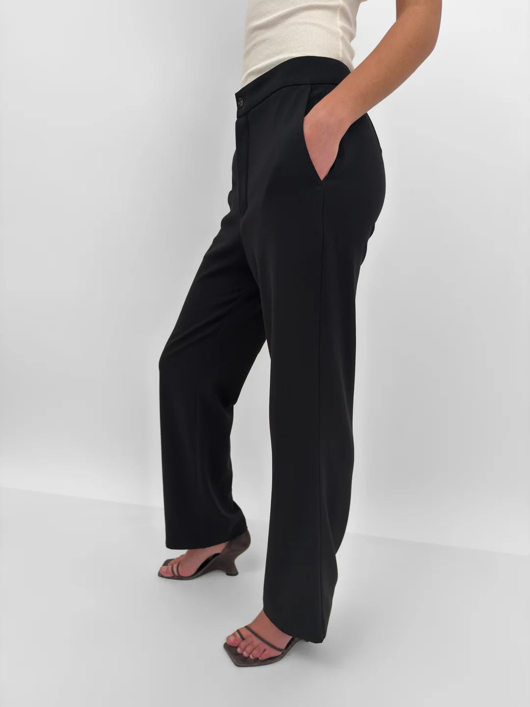 A model wearing black wide-leg trousers with side pockets, paired with a cream ribbed top and dark heeled sandals, shown from the waist down against a light gray studio background.