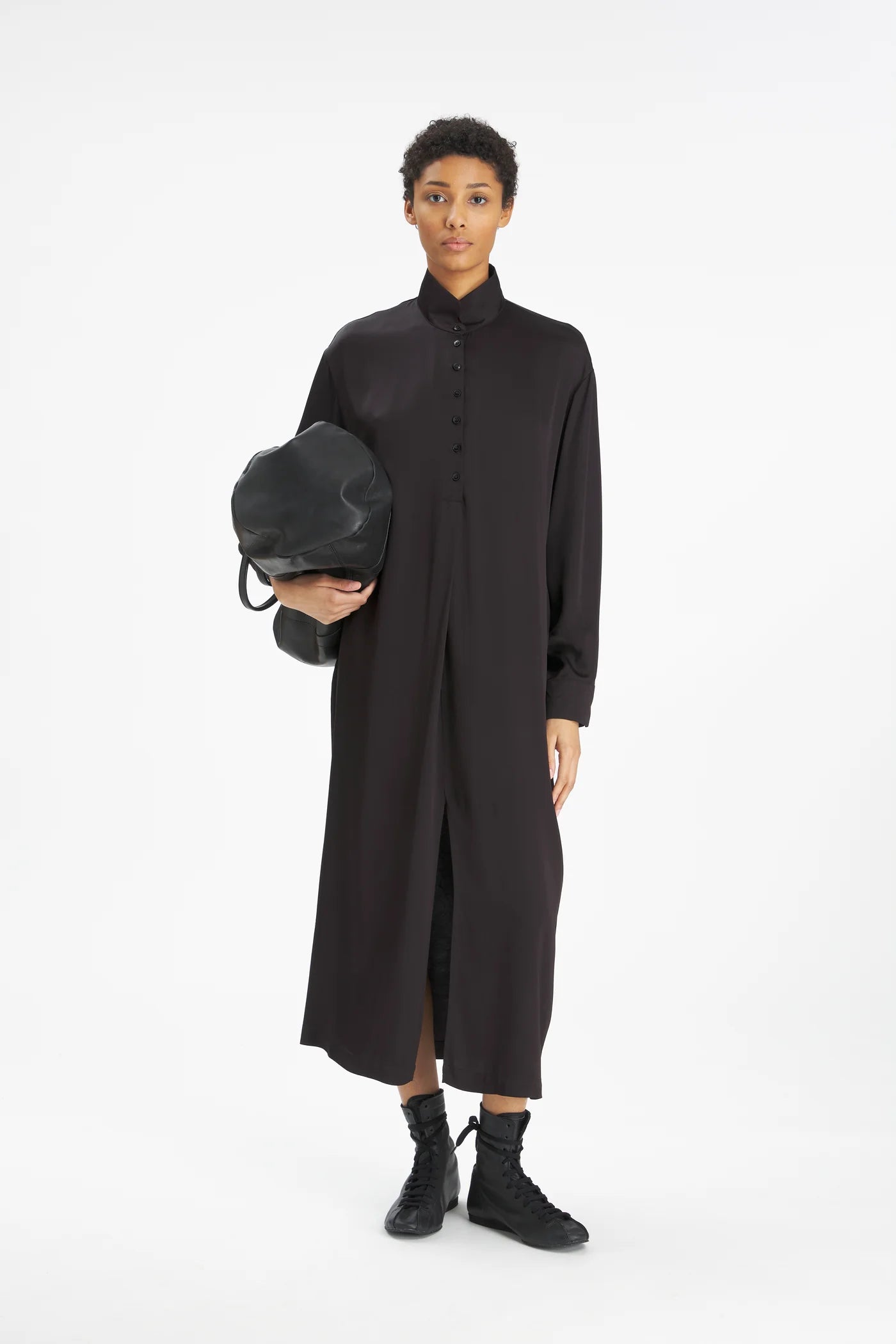 A model stands against a plain white studio background wearing a long black button-front dress with a high collar, black ankle boots, and holding a black bag.