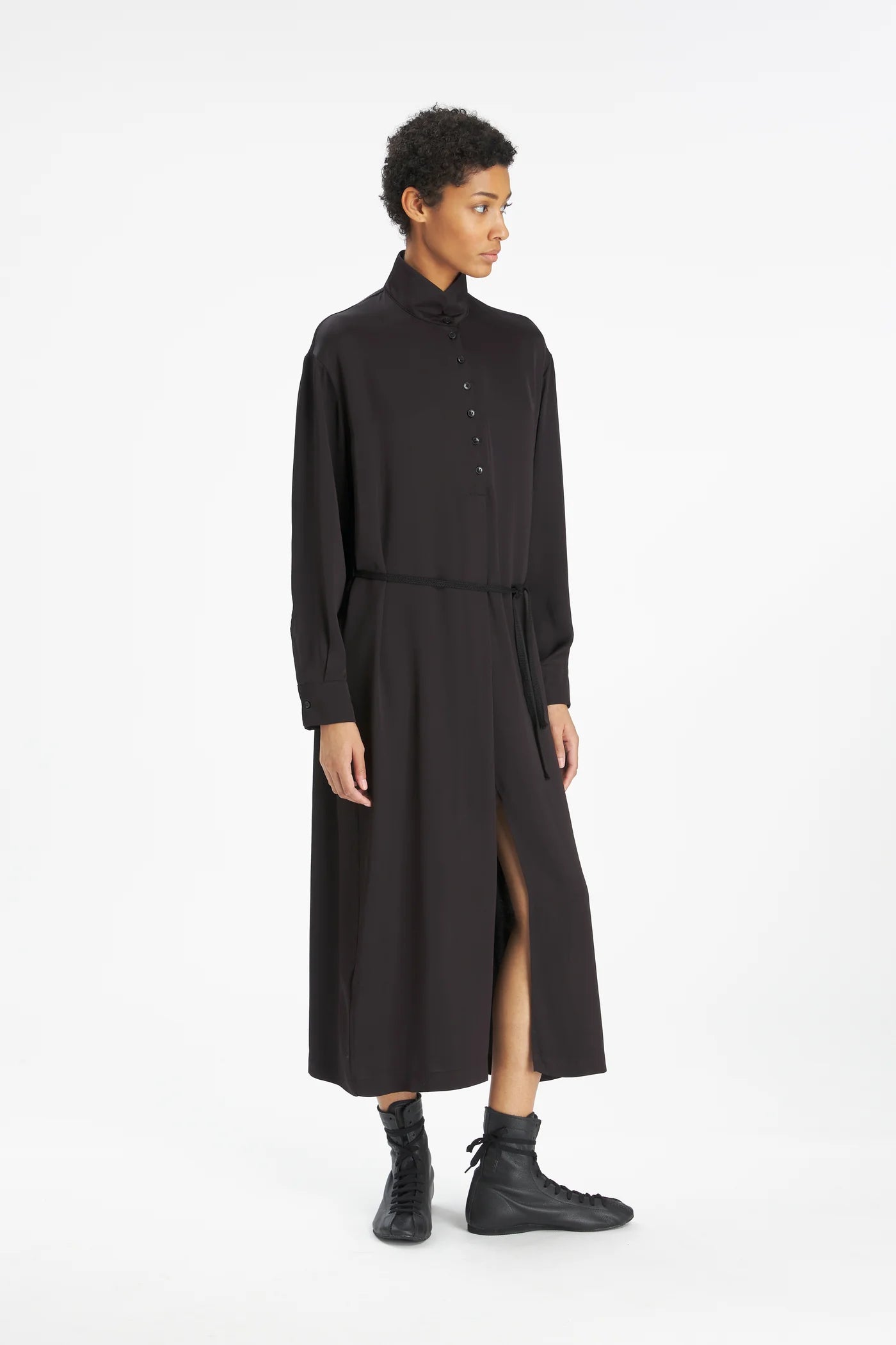A model wearing a black long-sleeve midi shirt dress with a high collar, buttoned front, and a side slit, paired with black boots, against a plain white studio background.