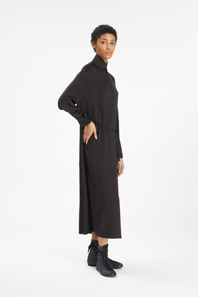 A model wearing a long-sleeved black turtleneck midi dress with a matching belt, paired with black lace-up ankle boots, photographed against a seamless white studio background.