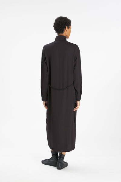 A model viewed from the back wearing a long-sleeved black high-neck robe or tunic dress with a tie at the waist, paired with black socks and shoes, against a plain white studio background.