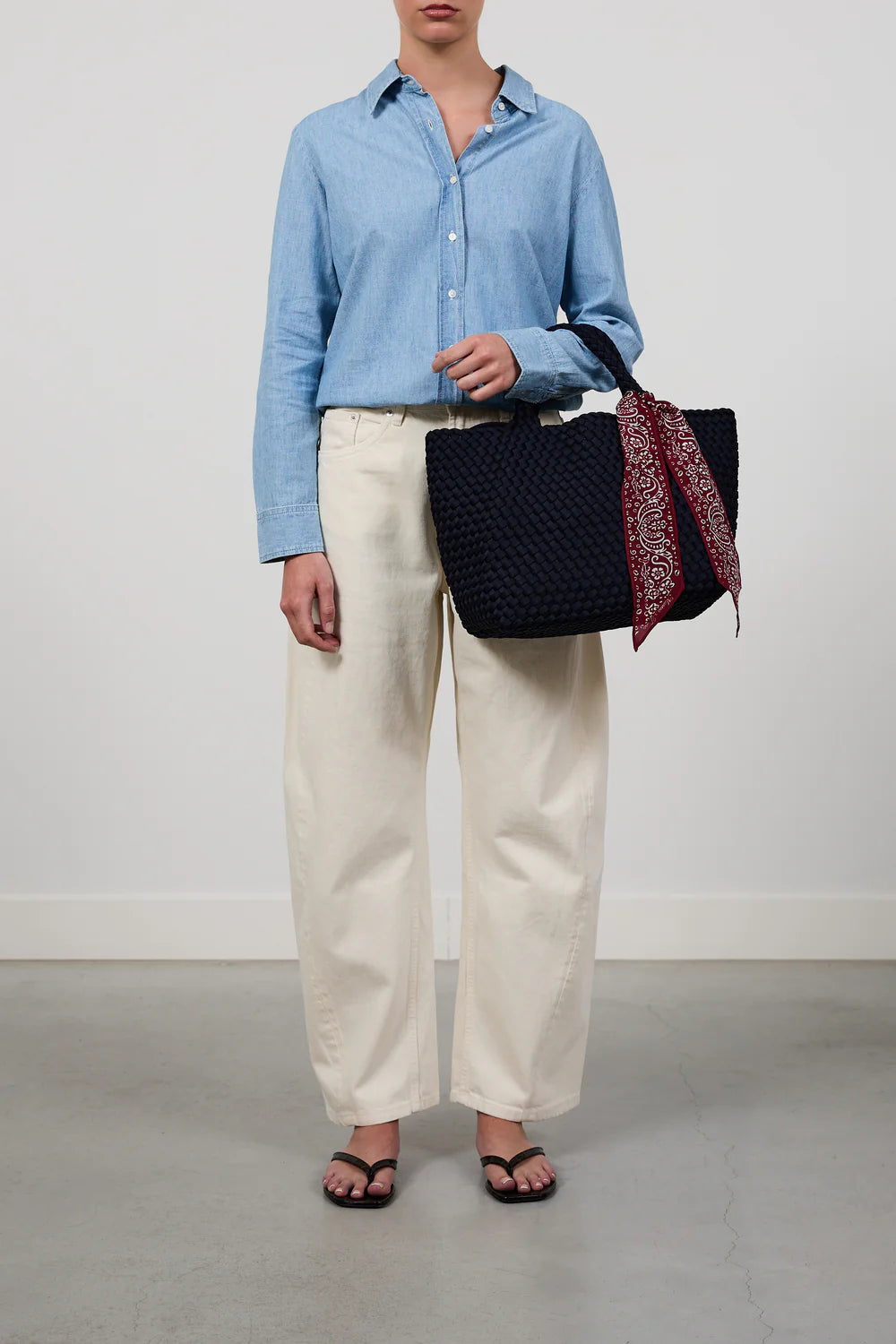 A model wearing a light blue button-down shirt, cream wide-leg trousers, and black sandals, holding a black woven tote bag with a red patterned scarf.