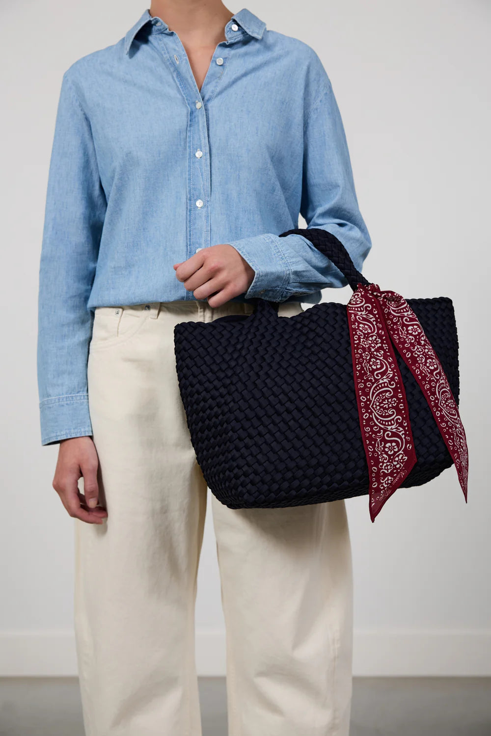 A person wearing a light blue button-up shirt and cream trousers holds a large woven black tote bag with a red paisley bandana tied to its handle, against a plain light gray wall.