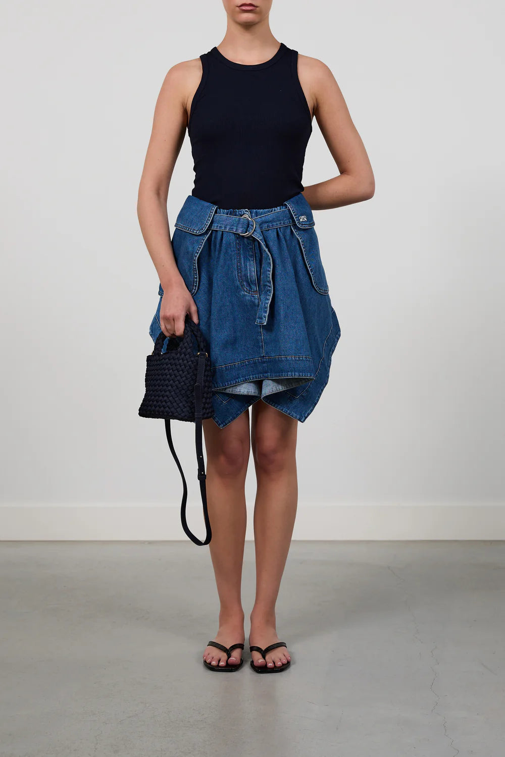 A model wearing a sleeveless black ribbed tank top, a blue denim skirt with a tie belt, and black sandals, holding a black woven handbag against a plain light gray studio background.