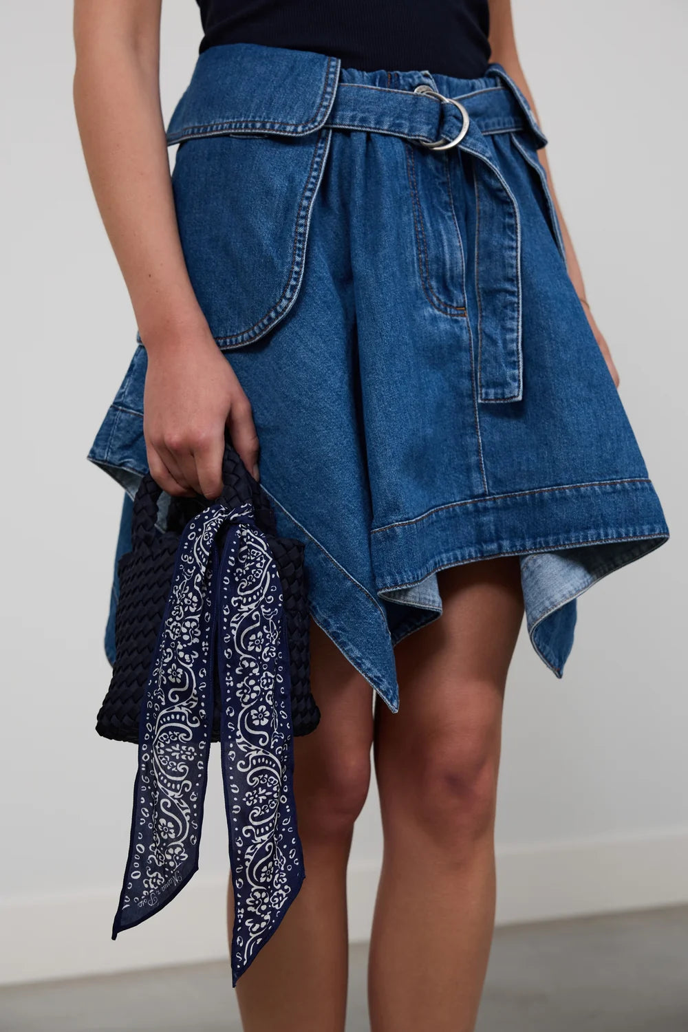 A model wearing a blue denim mini skirt with an asymmetrical hem, belt, and silver ring detail, holding a navy and white paisley bandana.