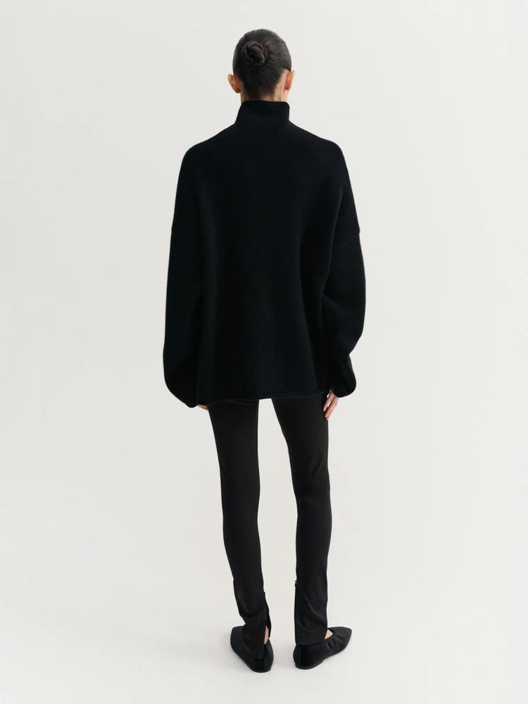 A model viewed from the back wearing a black oversized mock-neck sweater, black slim-fit pants, and black flats against a plain white studio background.