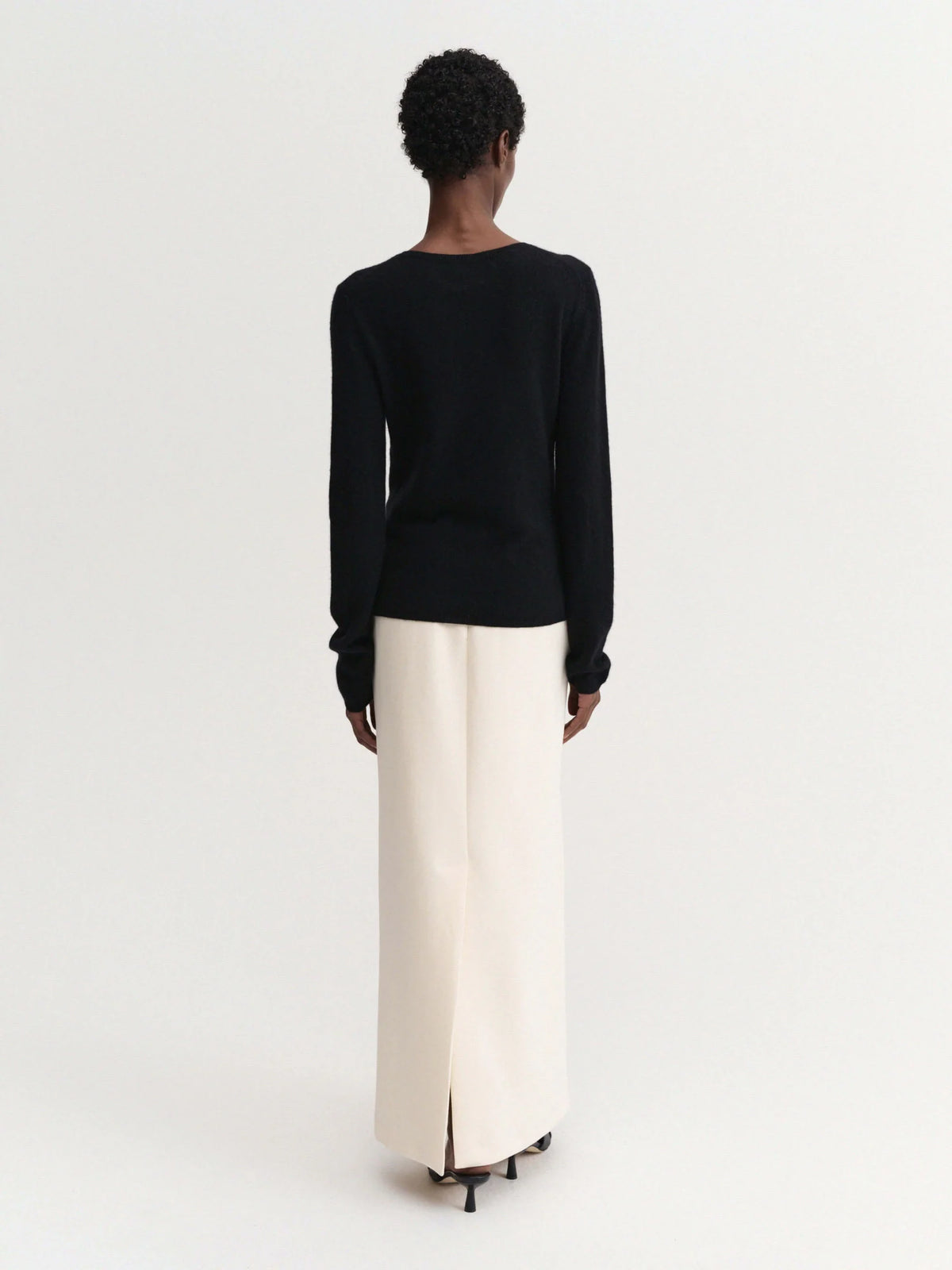 A model shown from the back wearing a black long-sleeve top and a cream-colored wide-leg skirt, photographed against a solid light gray studio background.