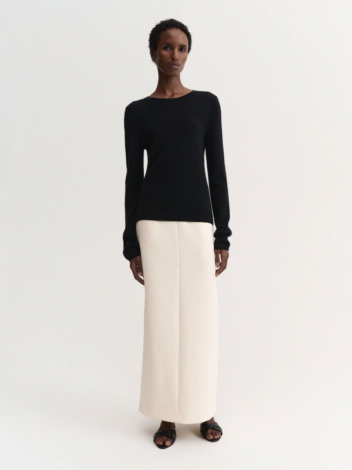 A model stands against a plain light background wearing a black long-sleeve knit top and a high-waisted, straight-leg cream skirt.
