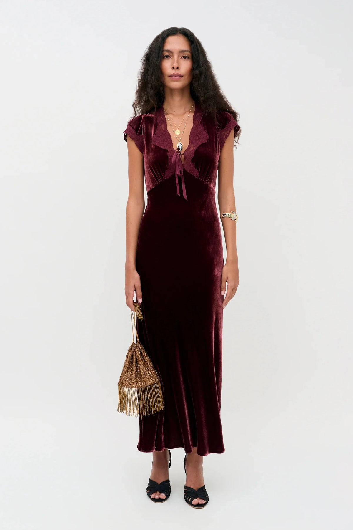 A woman modeling a floor-length burgundy velvet dress with a ruffled sweetheart neckline and cap sleeves, styled with a matching fringed handbag and black heeled sandals against a plain light gray studio background.