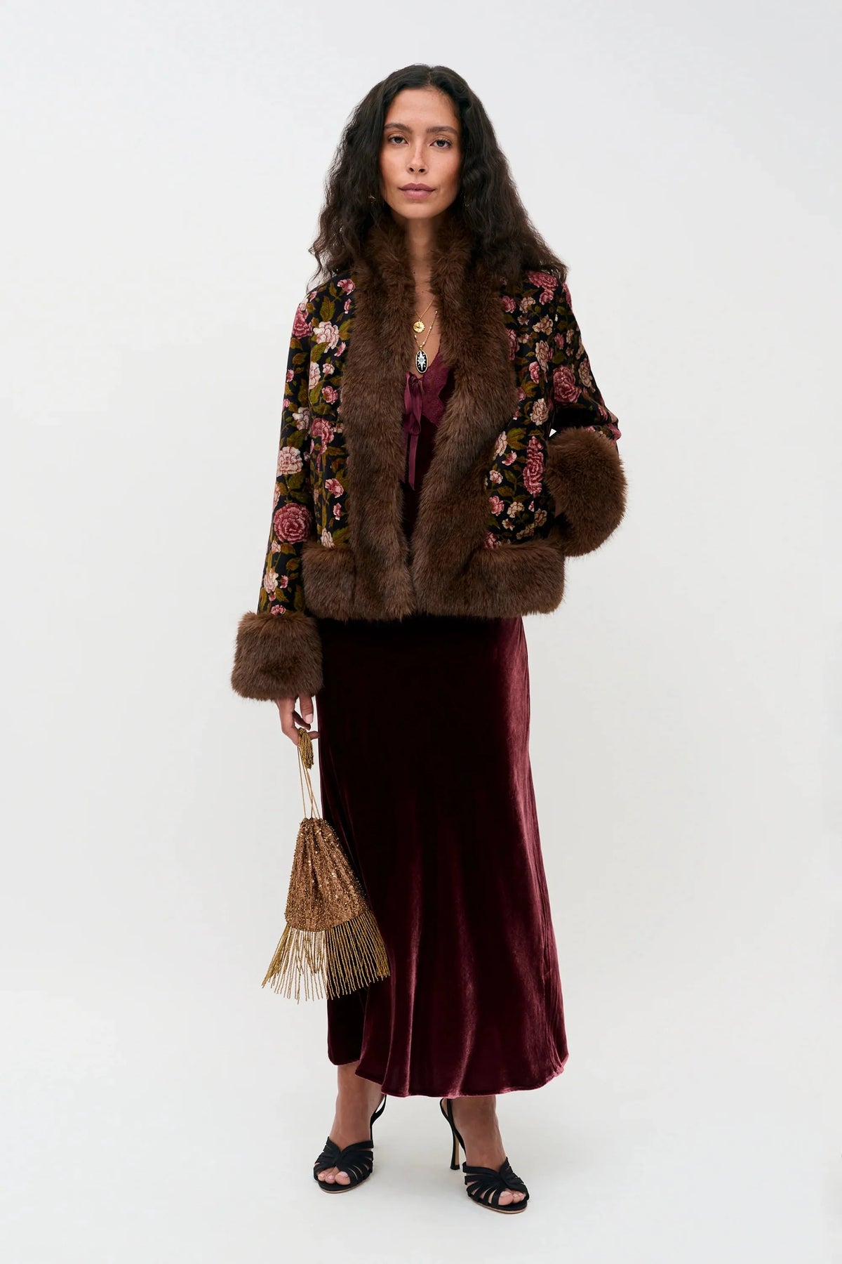 A model stands against a plain white studio background wearing a dark brown fur-trimmed jacket with colorful floral embroidery on the sleeves, a deep burgundy velvet midi skirt, and holding a small woven straw handbag.