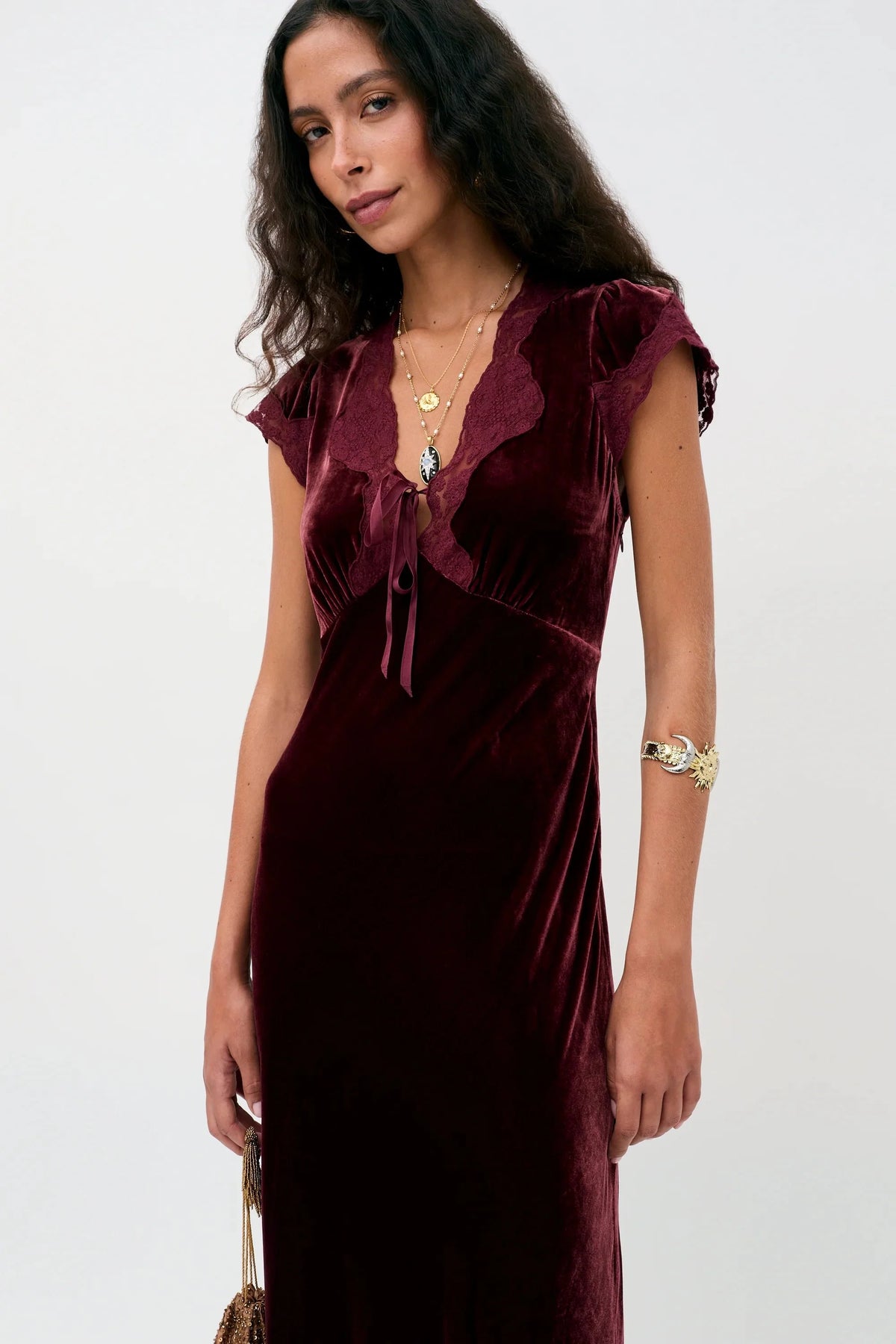 A woman modeling a deep burgundy velvet midi dress with cap sleeves, a ruffled V-neckline, and a front tie detail, set against a plain light gray background.