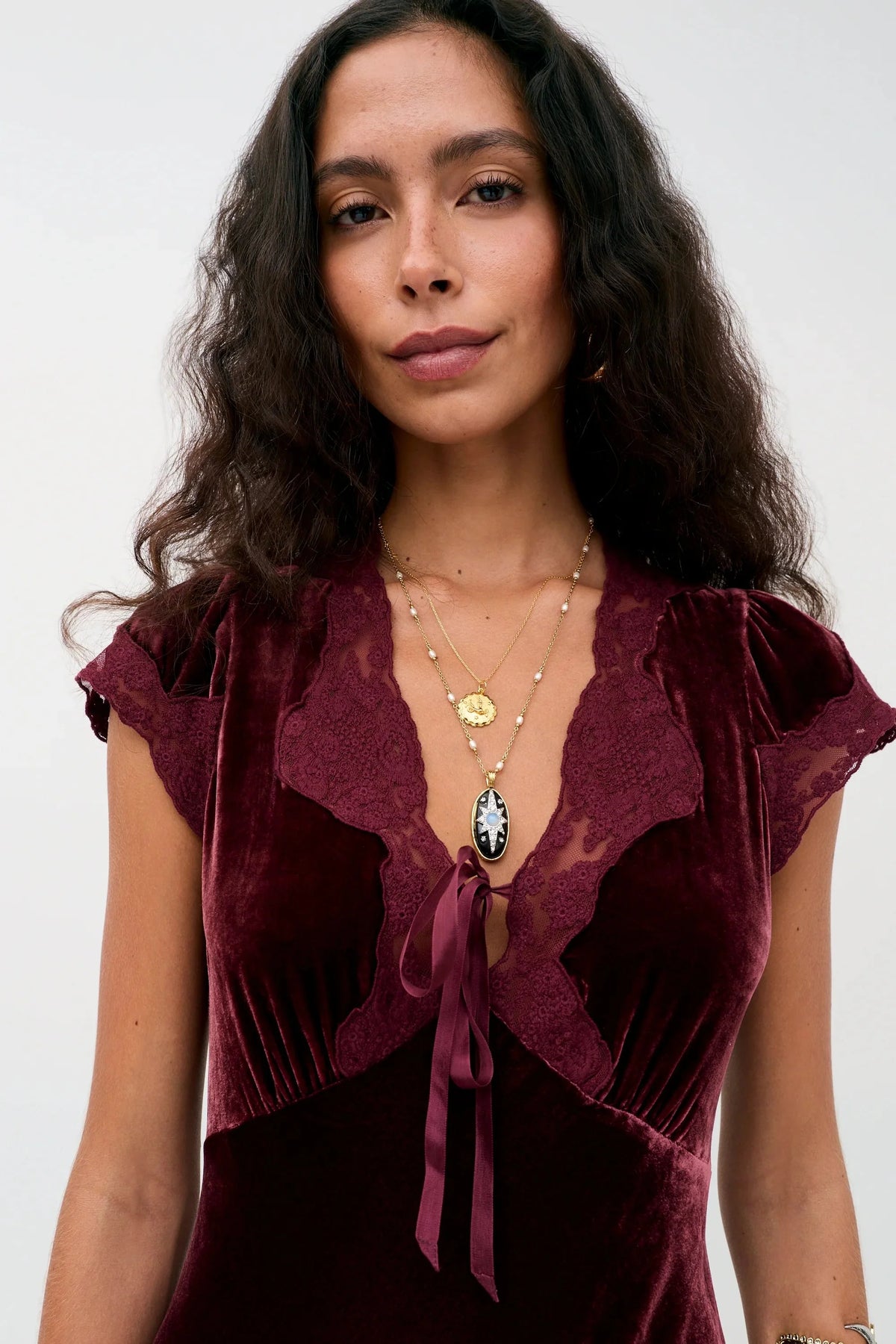 A woman wearing a deep burgundy velvet top with lace ruffle trim and a tie detail at the neckline, styled with layered gold necklaces against a plain light background.