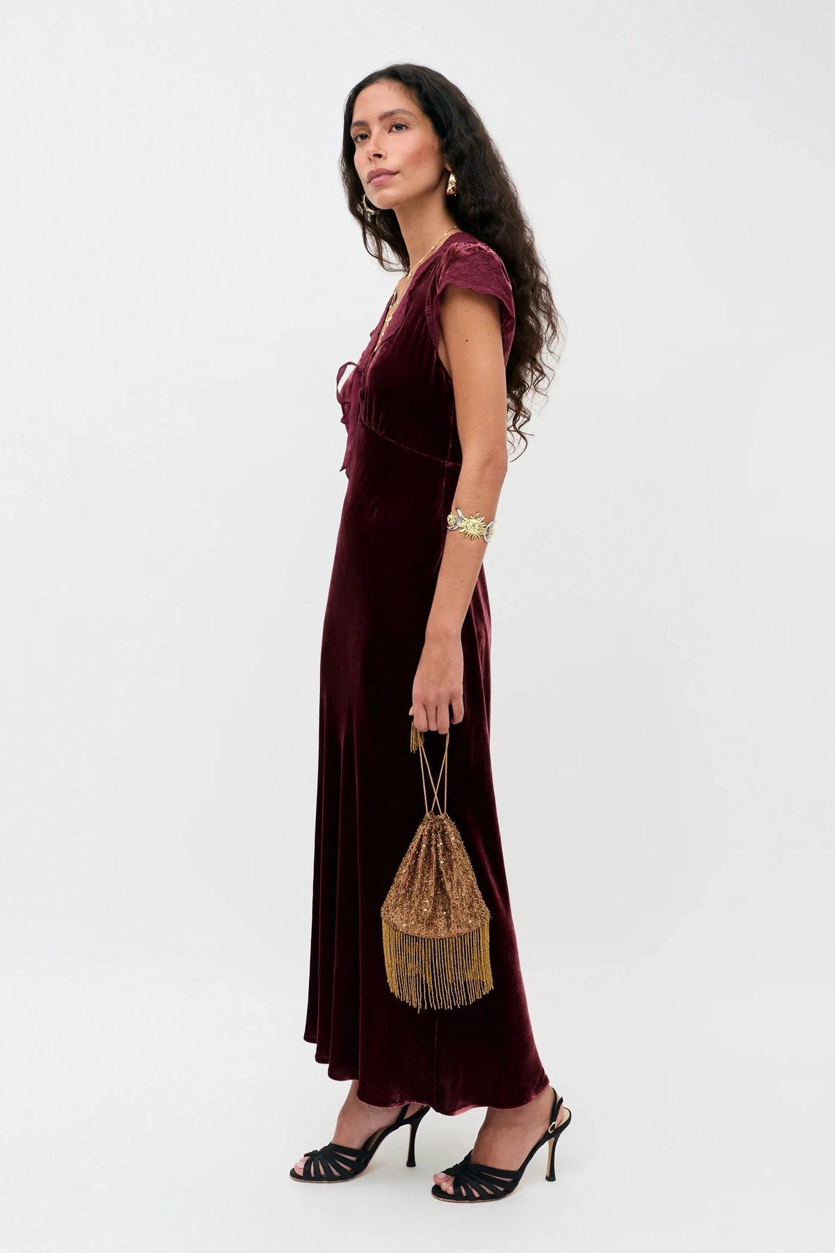 A woman modeling a floor-length burgundy velvet dress with cap sleeves, paired with black strappy heels and a small gold fringe bucket bag, against a plain white studio background.