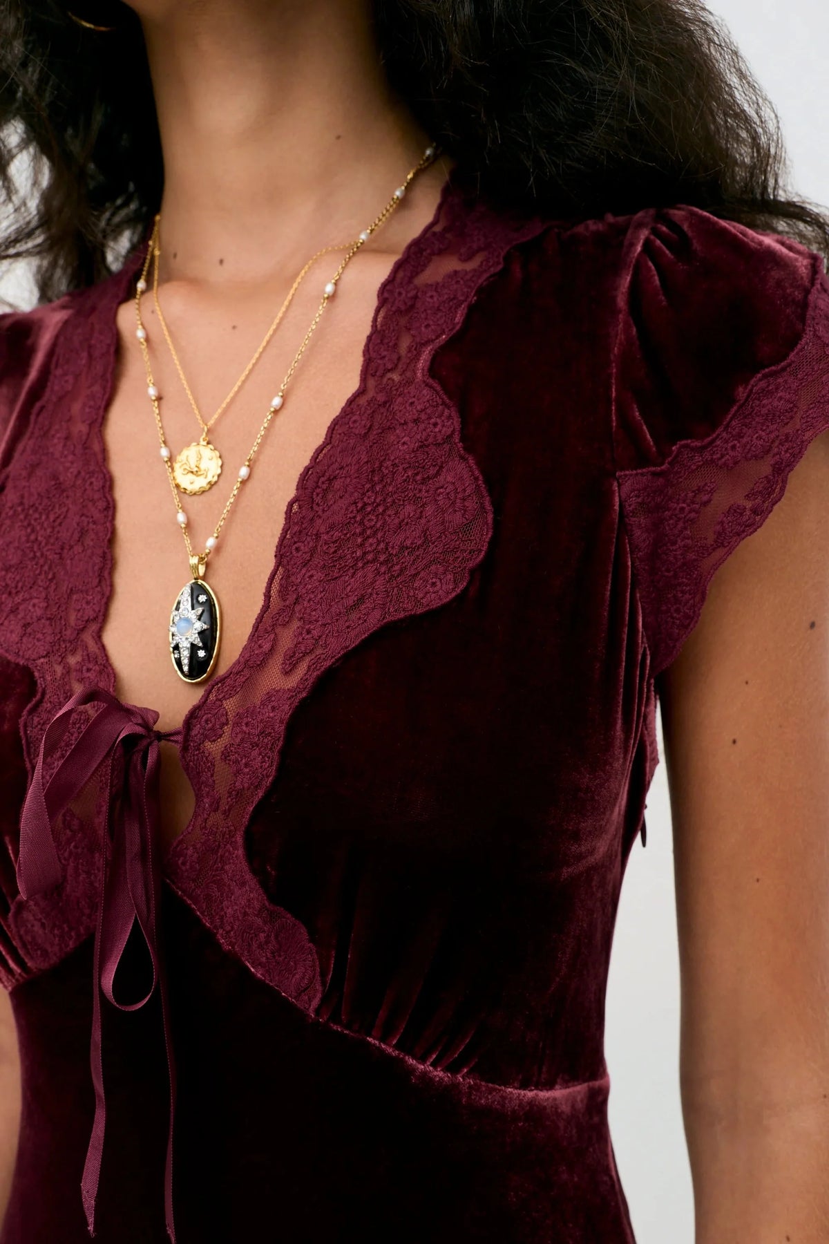 Close-up of a person wearing a deep burgundy velvet top with lace ruffle trim and a tie detail at the neckline, layered with gold necklaces.