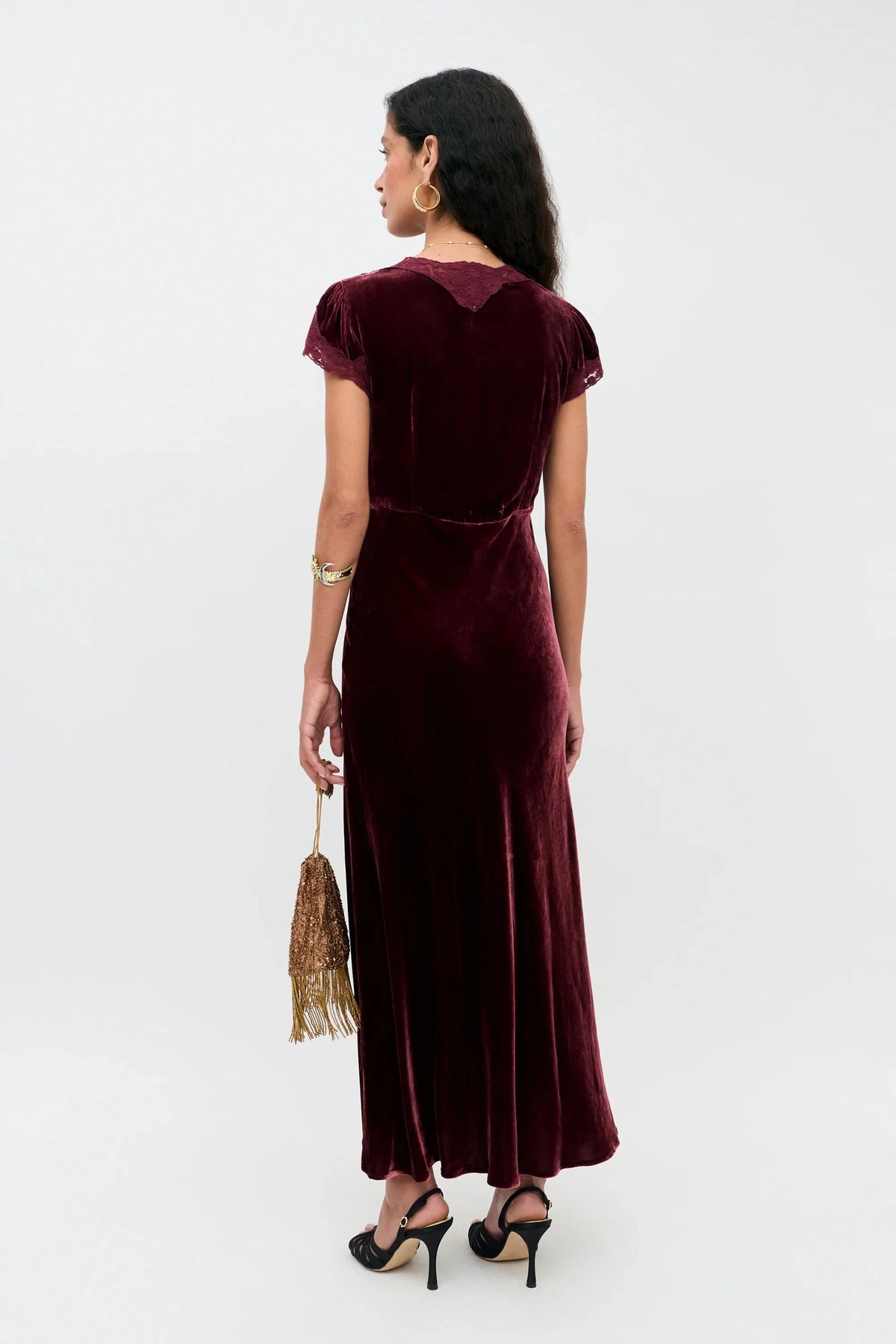 A woman stands with her back to the camera wearing a floor-length burgundy velvet dress with short cap sleeves and a matching belt at the waist. She is holding a small gold-fringed handbag and wearing black high heels against a plain white studio background.