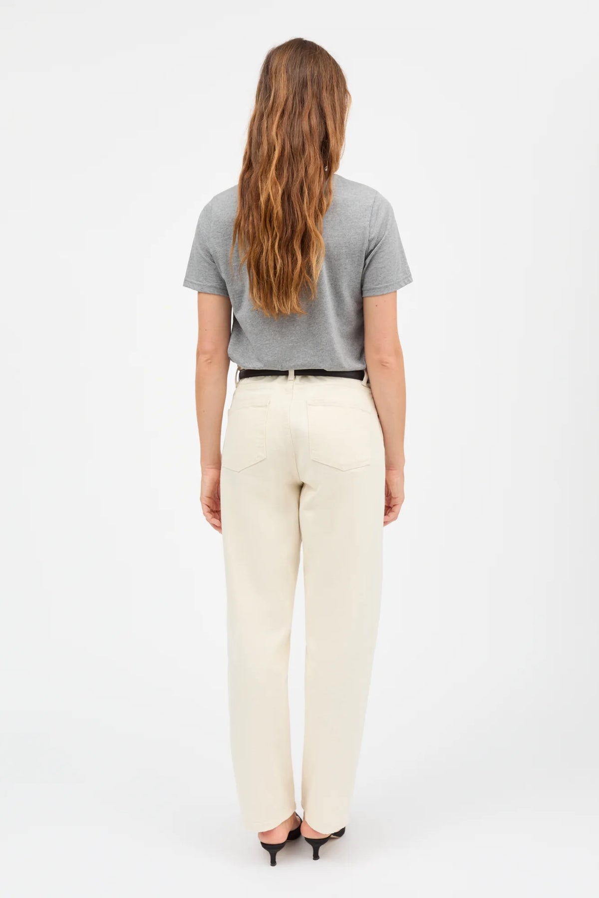 A woman viewed from behind wearing a gray short-sleeve t-shirt, cream-colored wide-leg trousers, a black belt, and black heels, set against a plain white studio background.