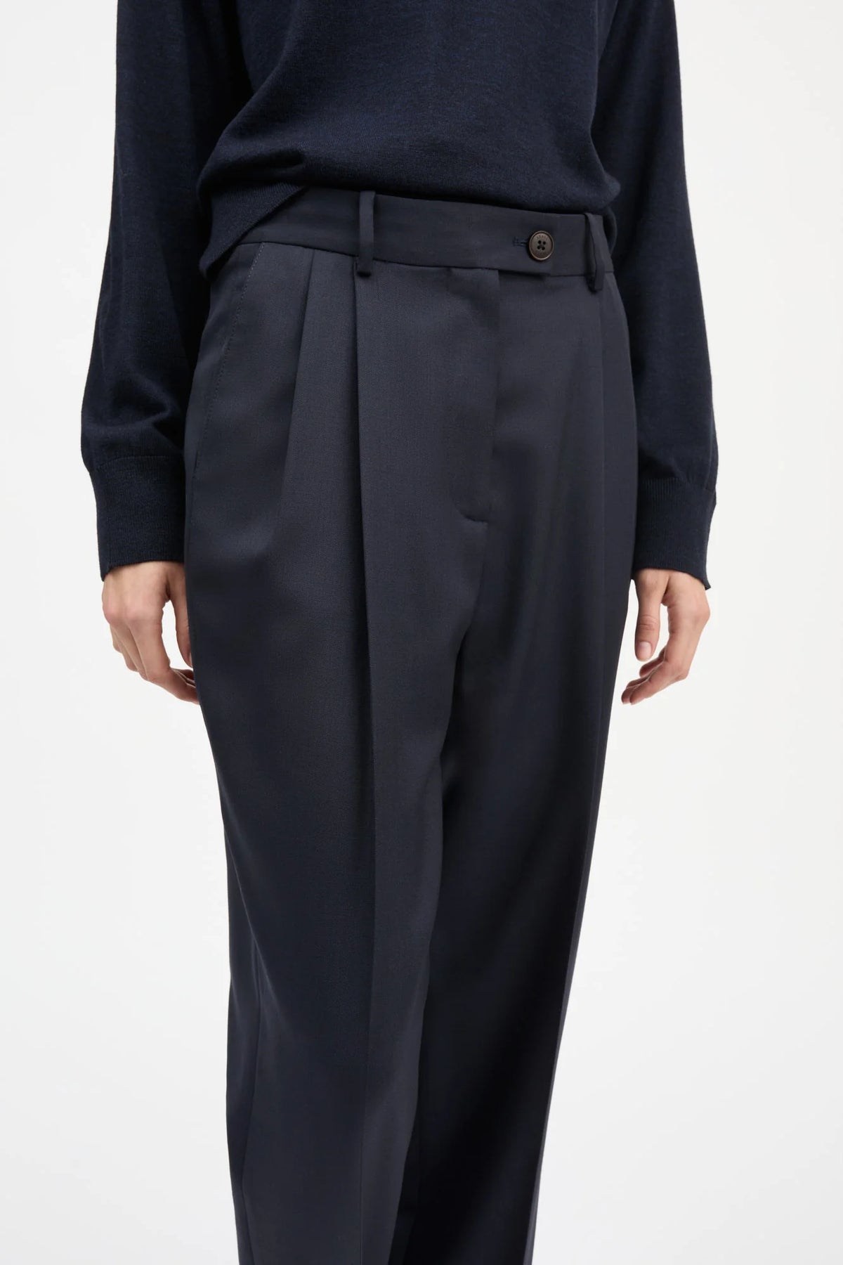 A close-up of a person wearing dark navy pleated trousers with a matching belt and a dark navy long-sleeve top, shown against a plain light gray studio background.