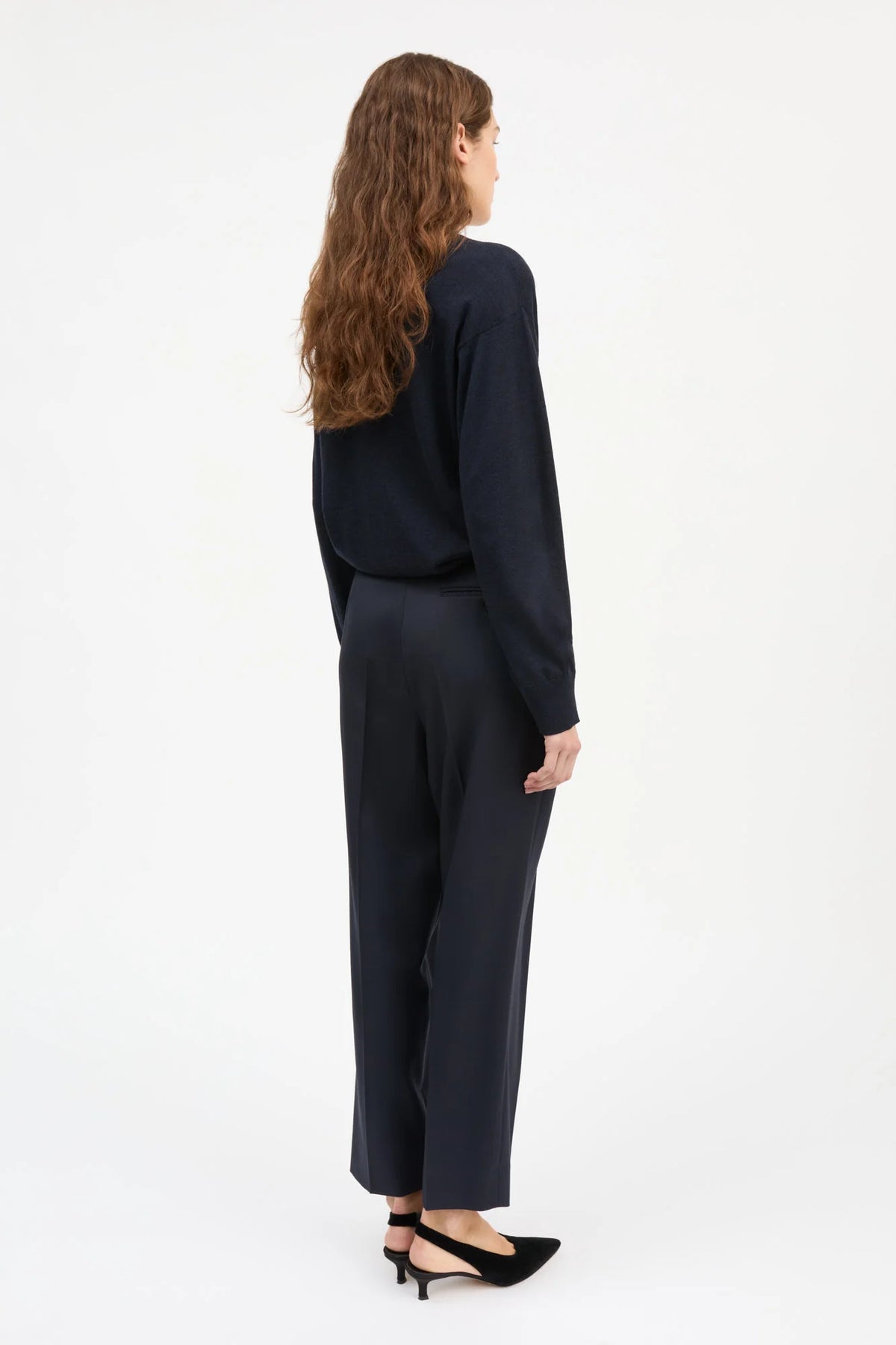 A woman with long wavy brown hair is shown from the back wearing a dark navy long-sleeve top and matching dark navy wide-leg trousers, paired with black pointed-toe heels, against a plain white studio background.