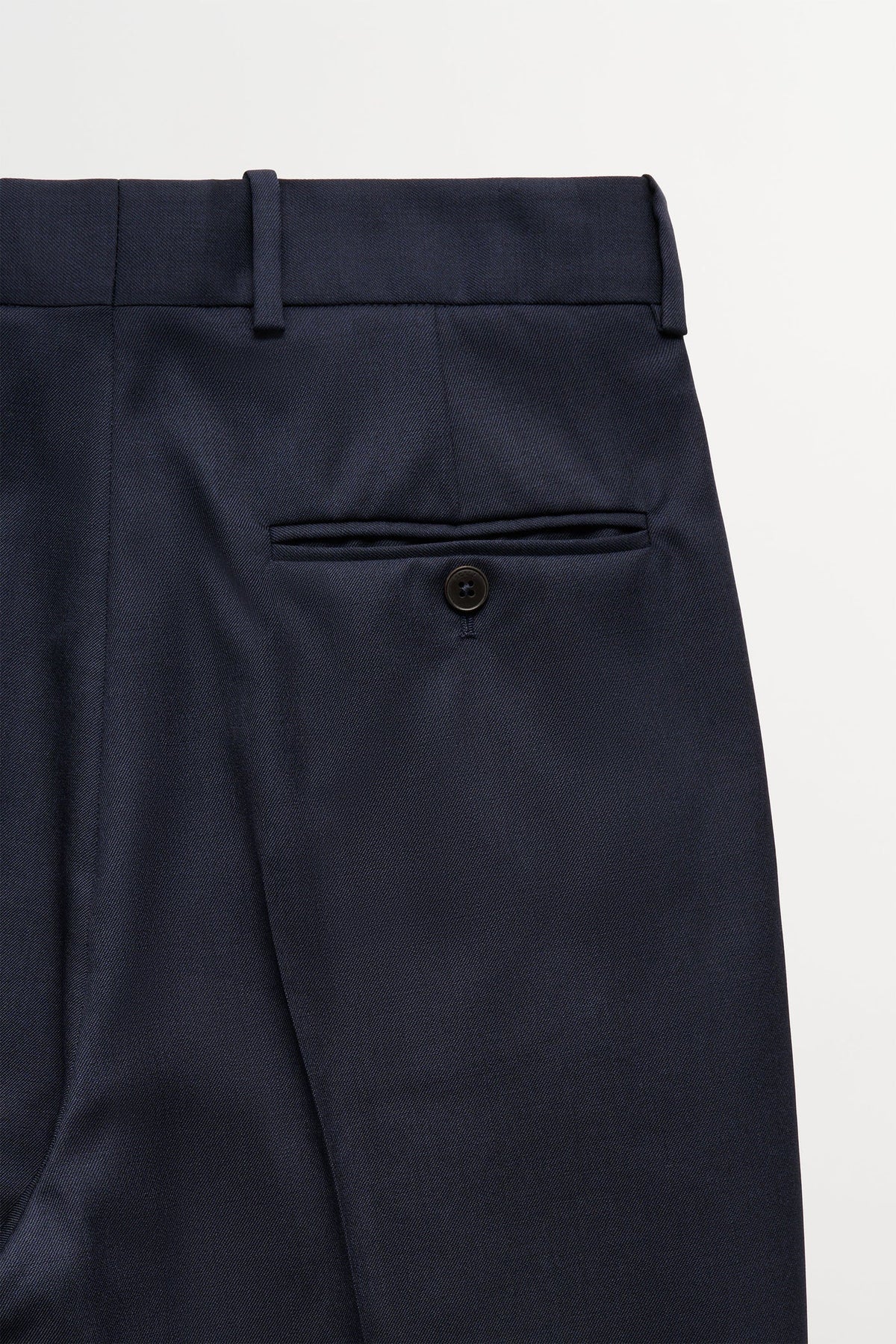 Close-up of navy blue trousers showing the waistband, belt loops, a side welt pocket with a button, and the fabric texture against a clean white background.