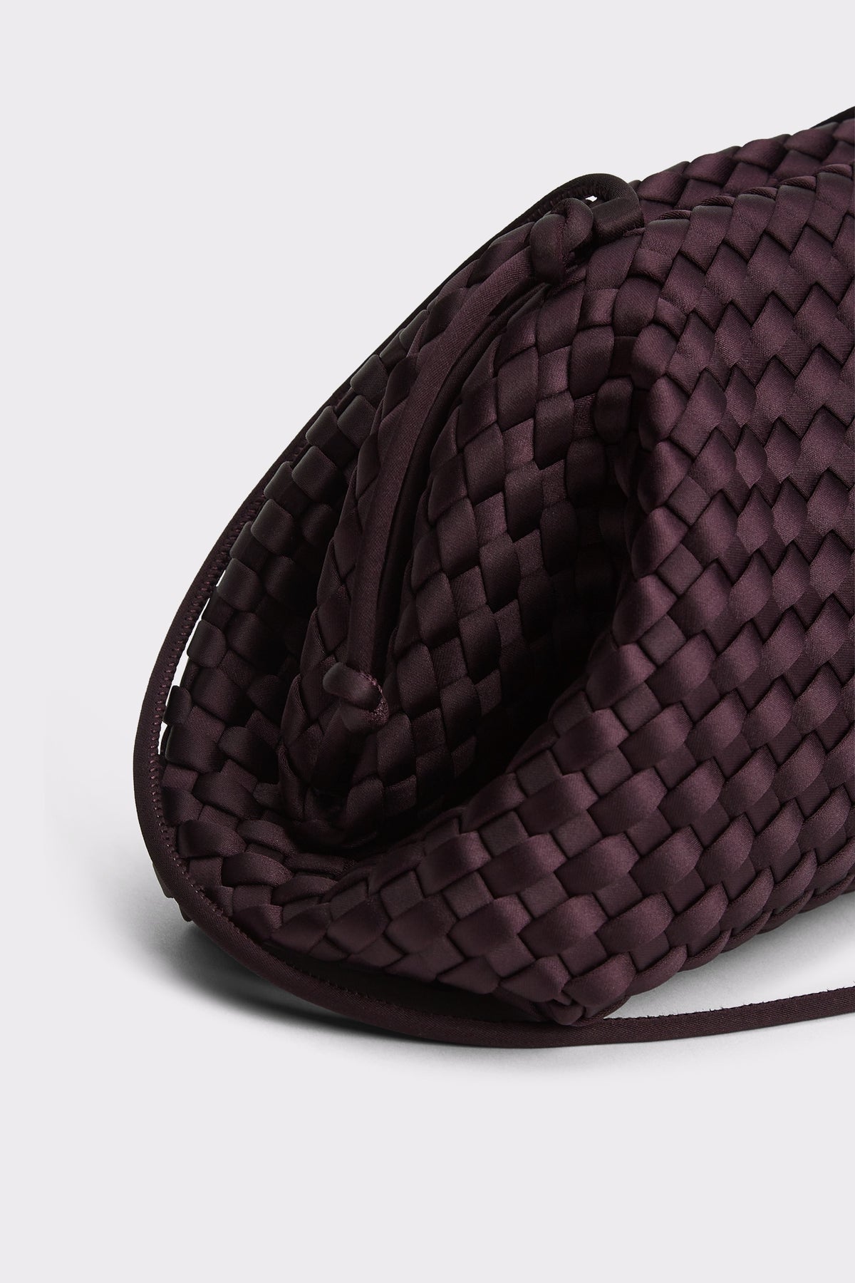 A close-up of a dark plum purple woven leather clutch bag, showing the detailed basketweave texture and curved silhouette against a light gray background.