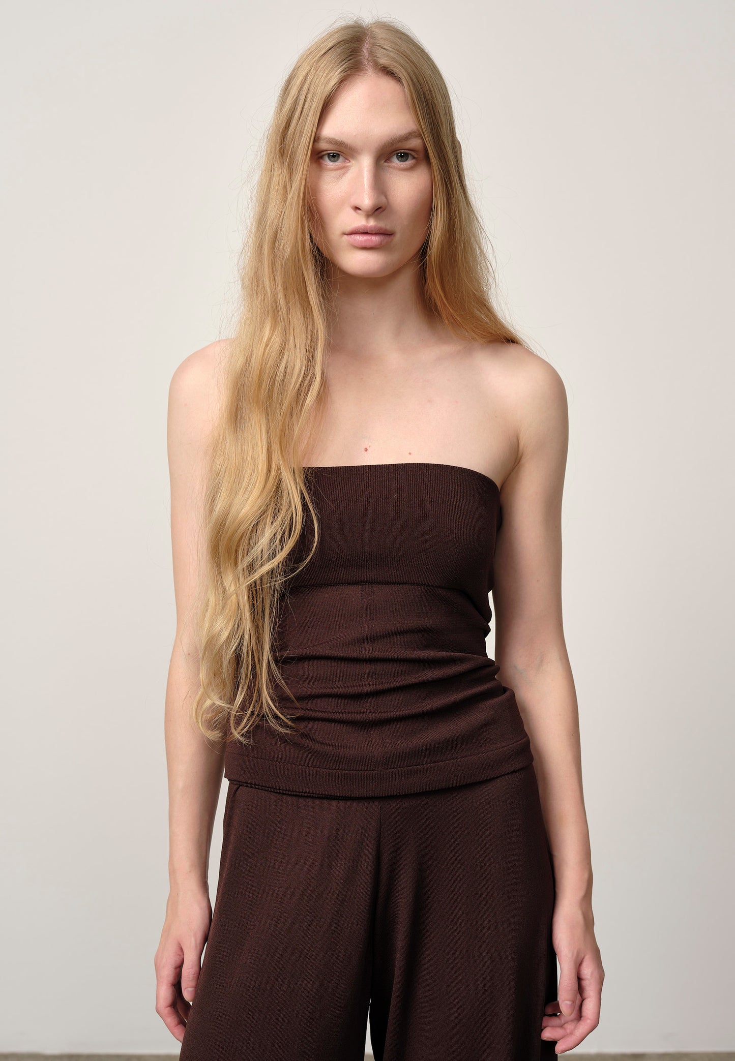 Woman wearing a brown strapless top and wide-leg pants against a plain background