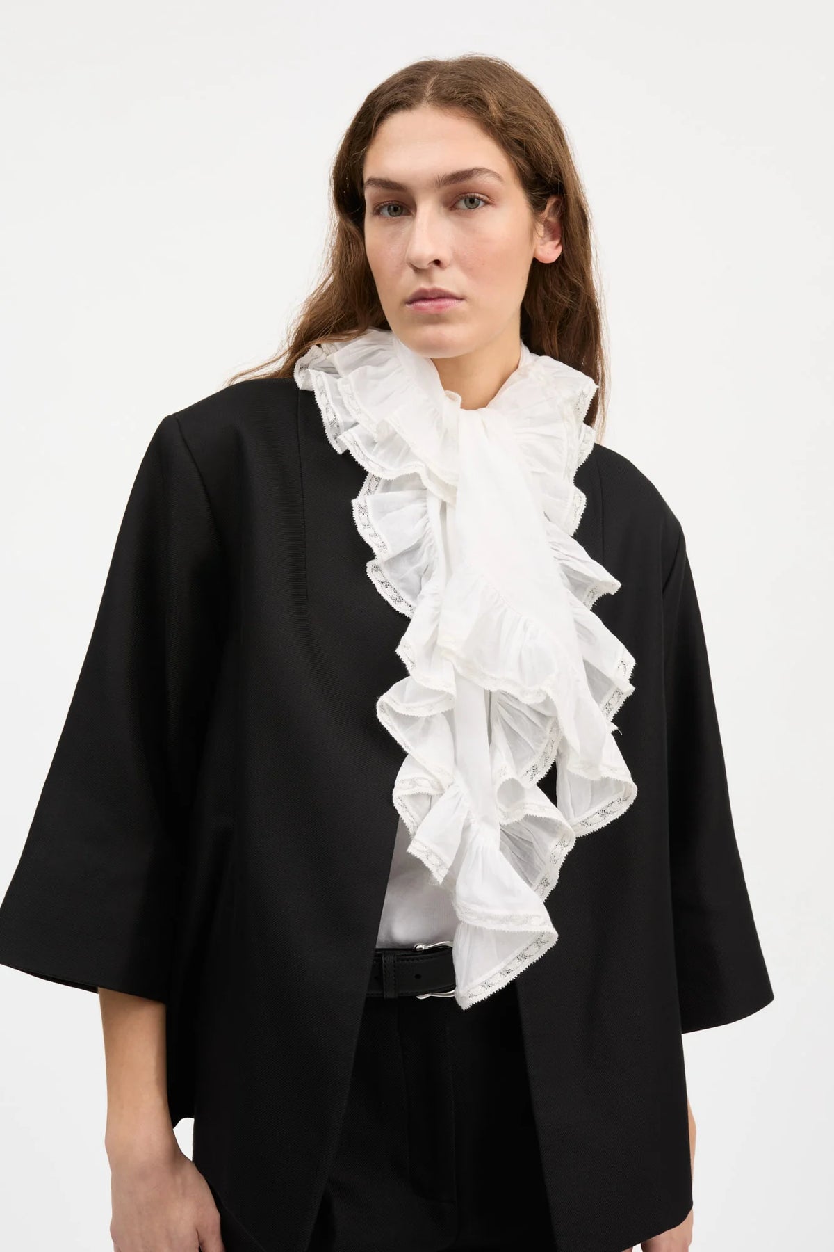 A model wearing a black draped cape-style jacket over a white ruffled blouse, photographed against a plain light gray studio background.