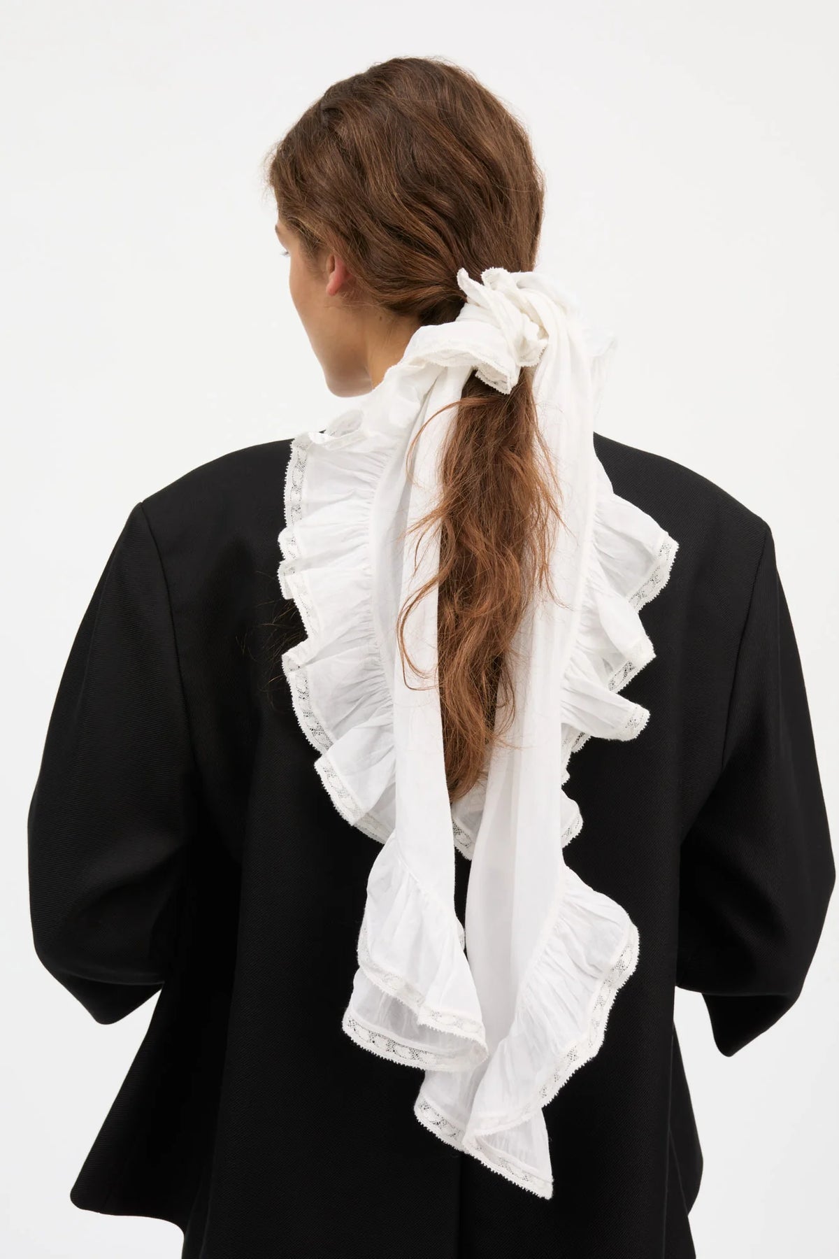 A woman viewed from behind wearing a voluminous white ruffled hair accessory tied in a ponytail, paired with a black top, against a plain white studio background.