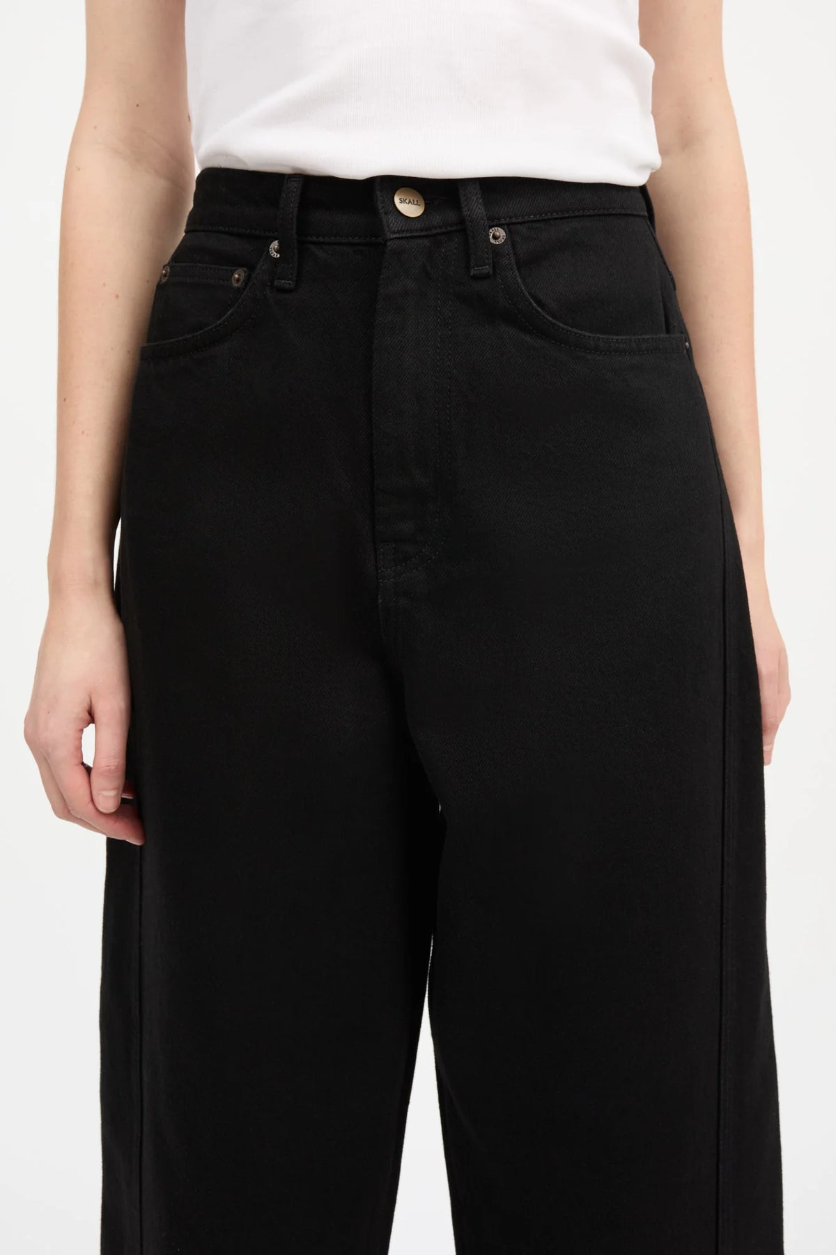 A close-up of a model wearing black wide-leg jeans with a white top, showing the front pockets, waistband, and side seam details against a plain light background.