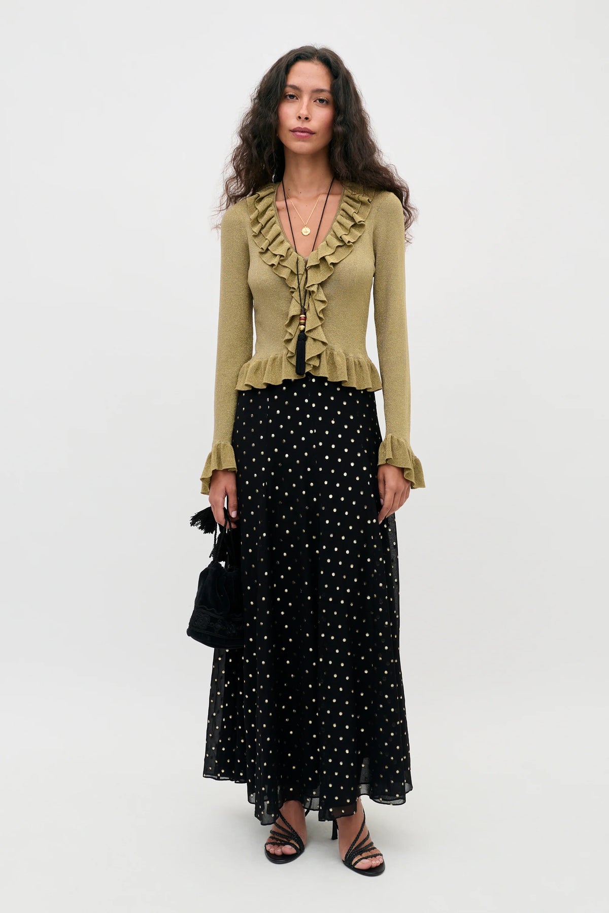 A model wearing a long black skirt with a gold polka-dot pattern, paired with a beige ruffled top and black strappy heels, holding a small black handbag