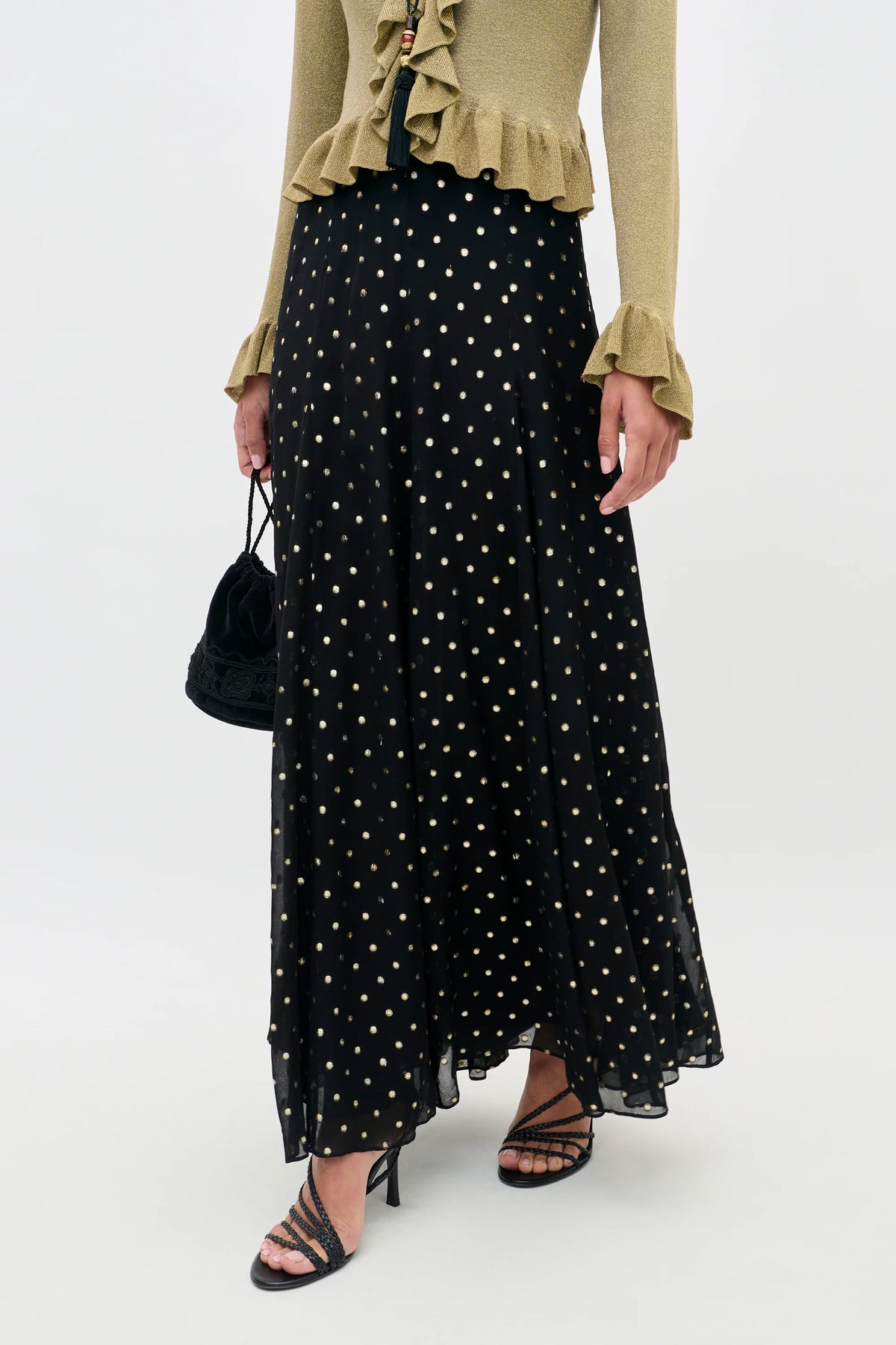 A model wearing a long black skirt with a gold polka-dot pattern, paired with a beige ruffled top and black strappy heels, holding a small black handbag against a light gray studio background.