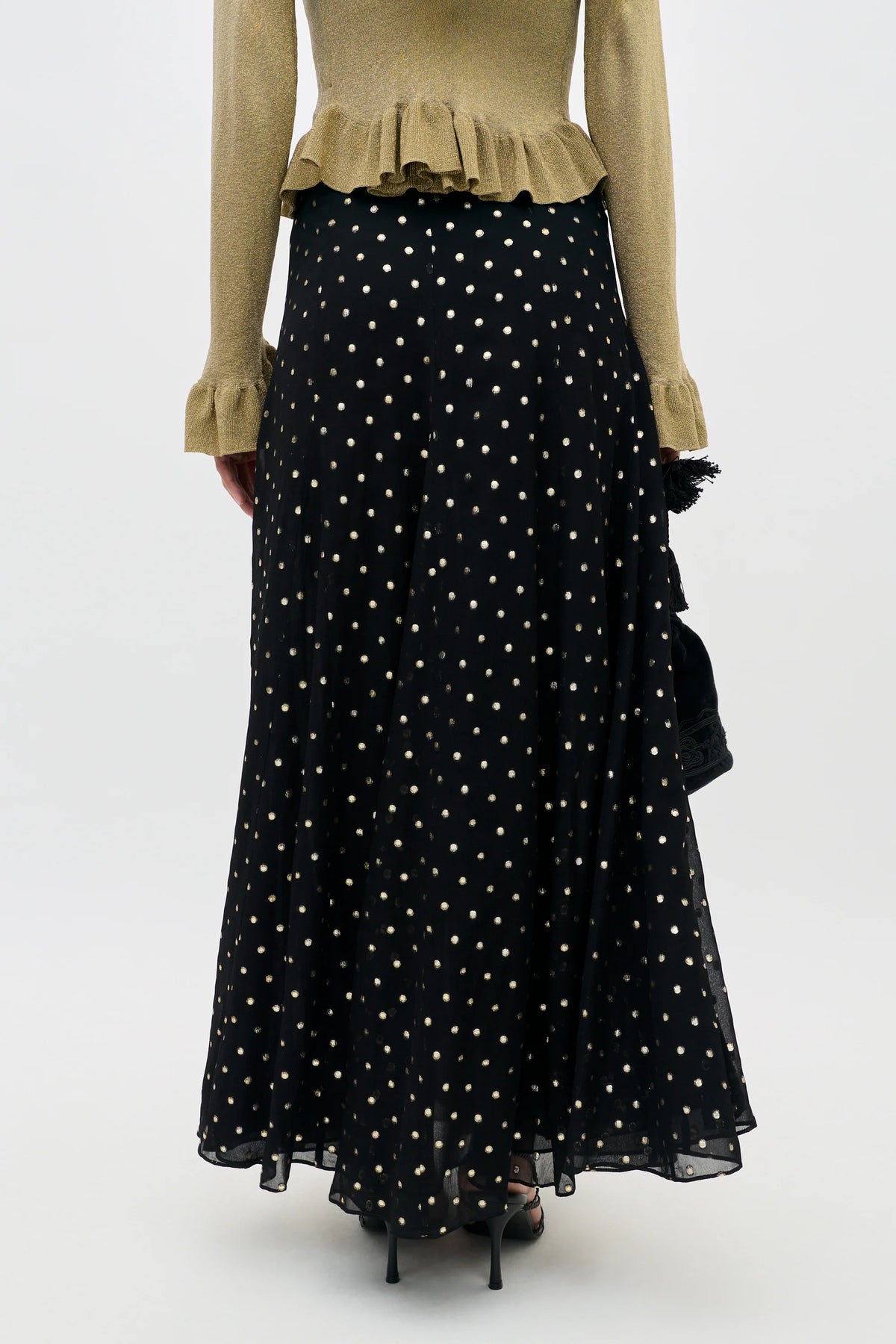 A model wearing a long black skirt with a gold polka-dot pattern, paired with a beige ruffled top and black strappy heels, holding a small black handbag