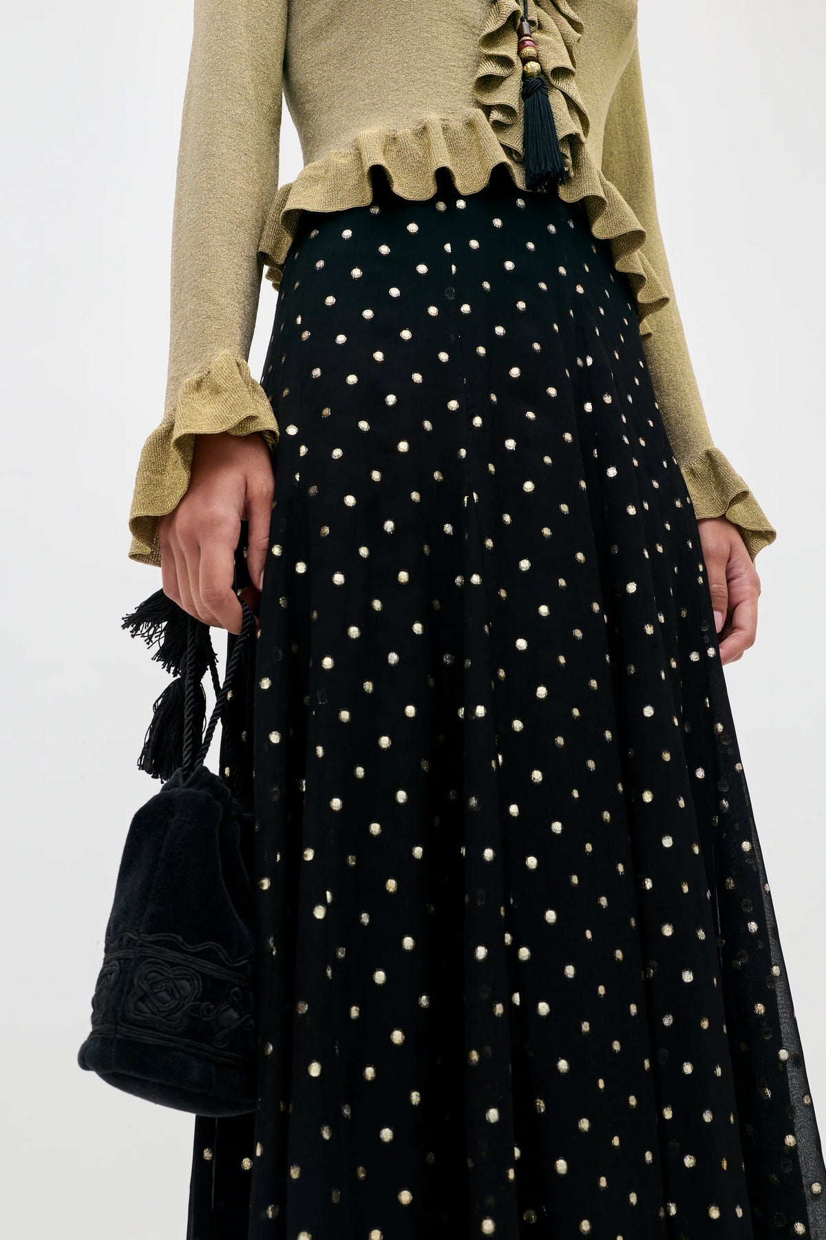 A model wearing a black skirt with gold polka dots and a beige ruffled top, holding a black tassel bucket bag against a plain white background.